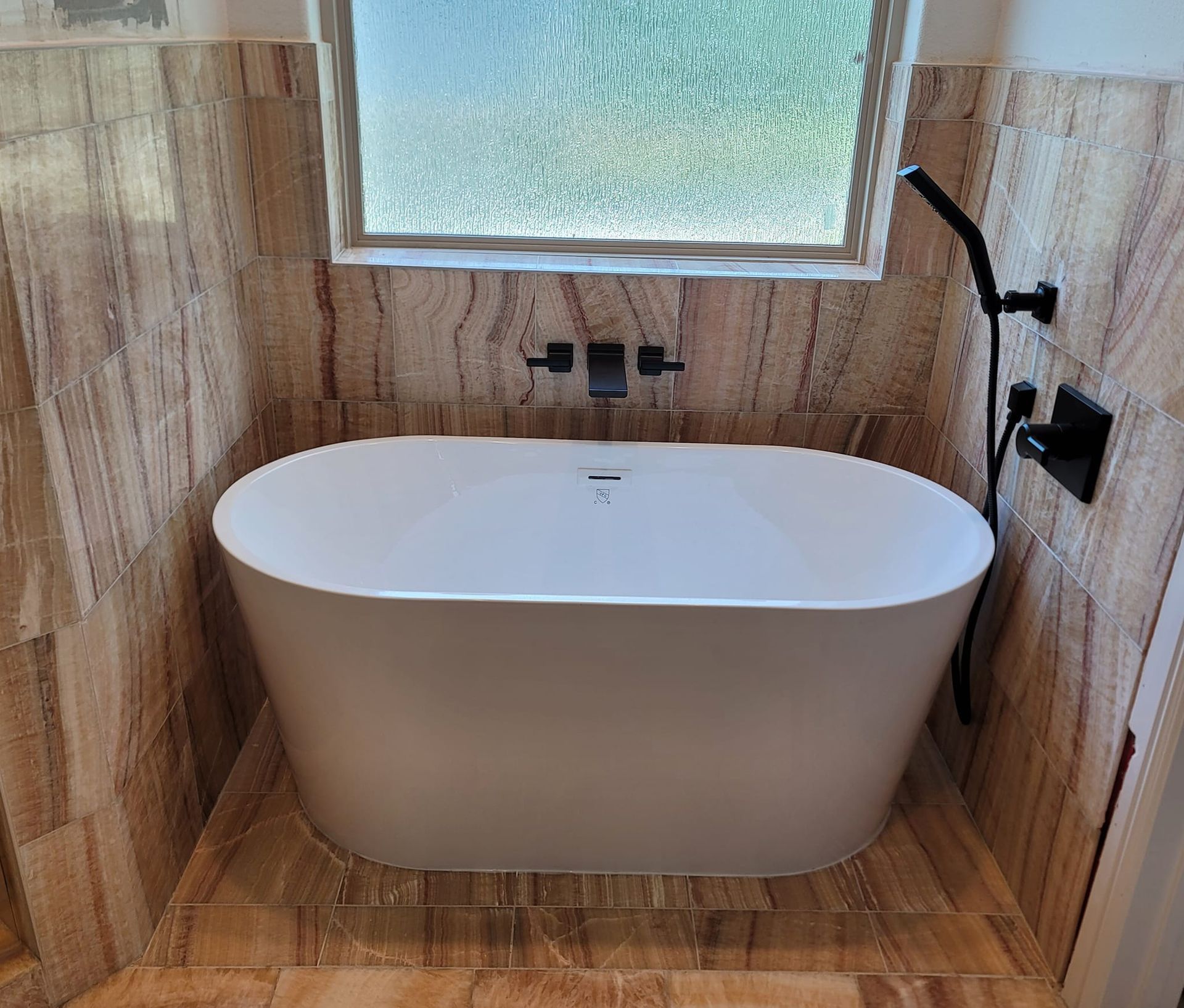 Freestanding white bathtub in a bathroom, with textured tile walls, black faucet and shower head.