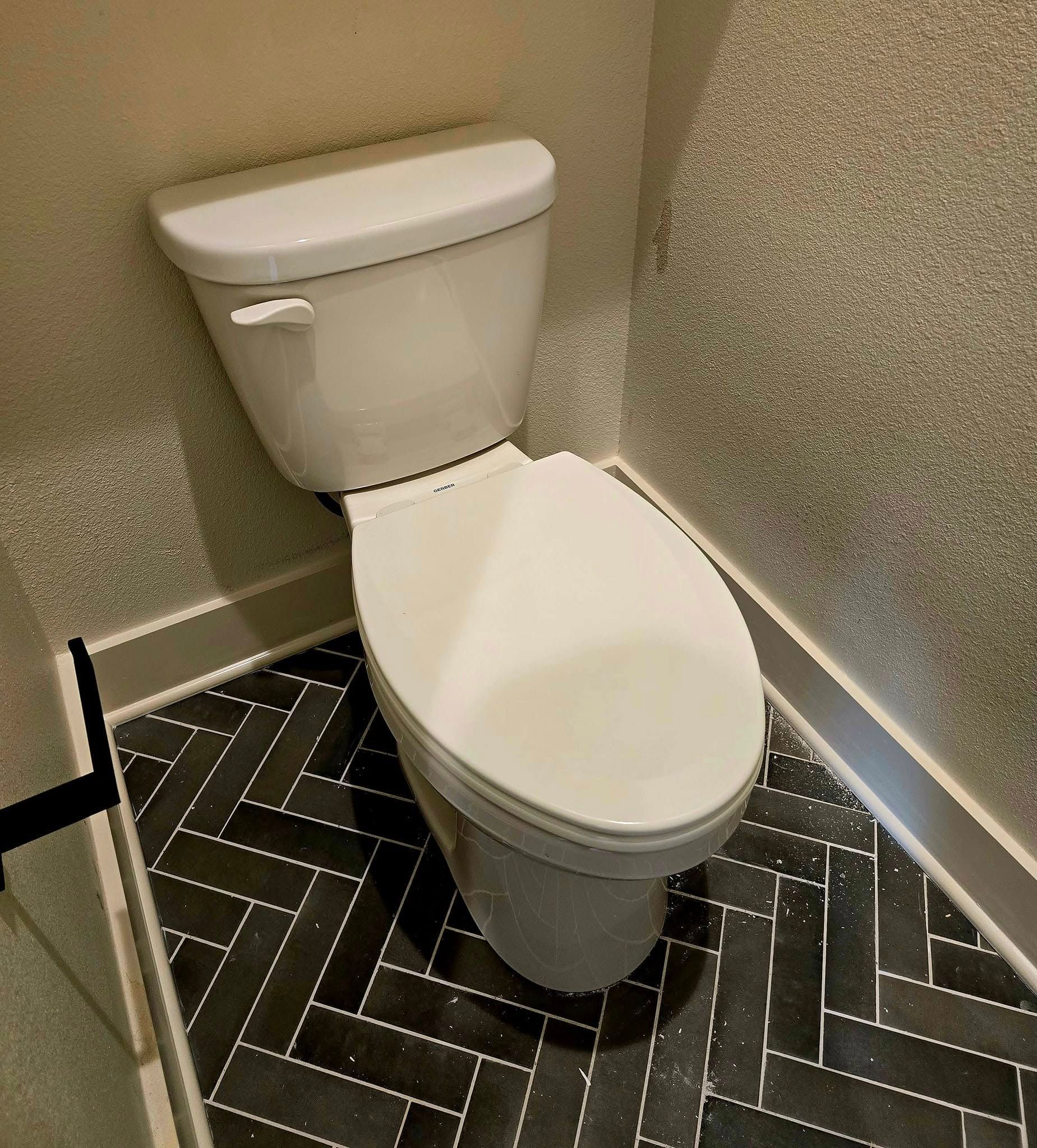 Toilet in a bathroom with dark, herringbone-patterned floor tiles and gray walls.