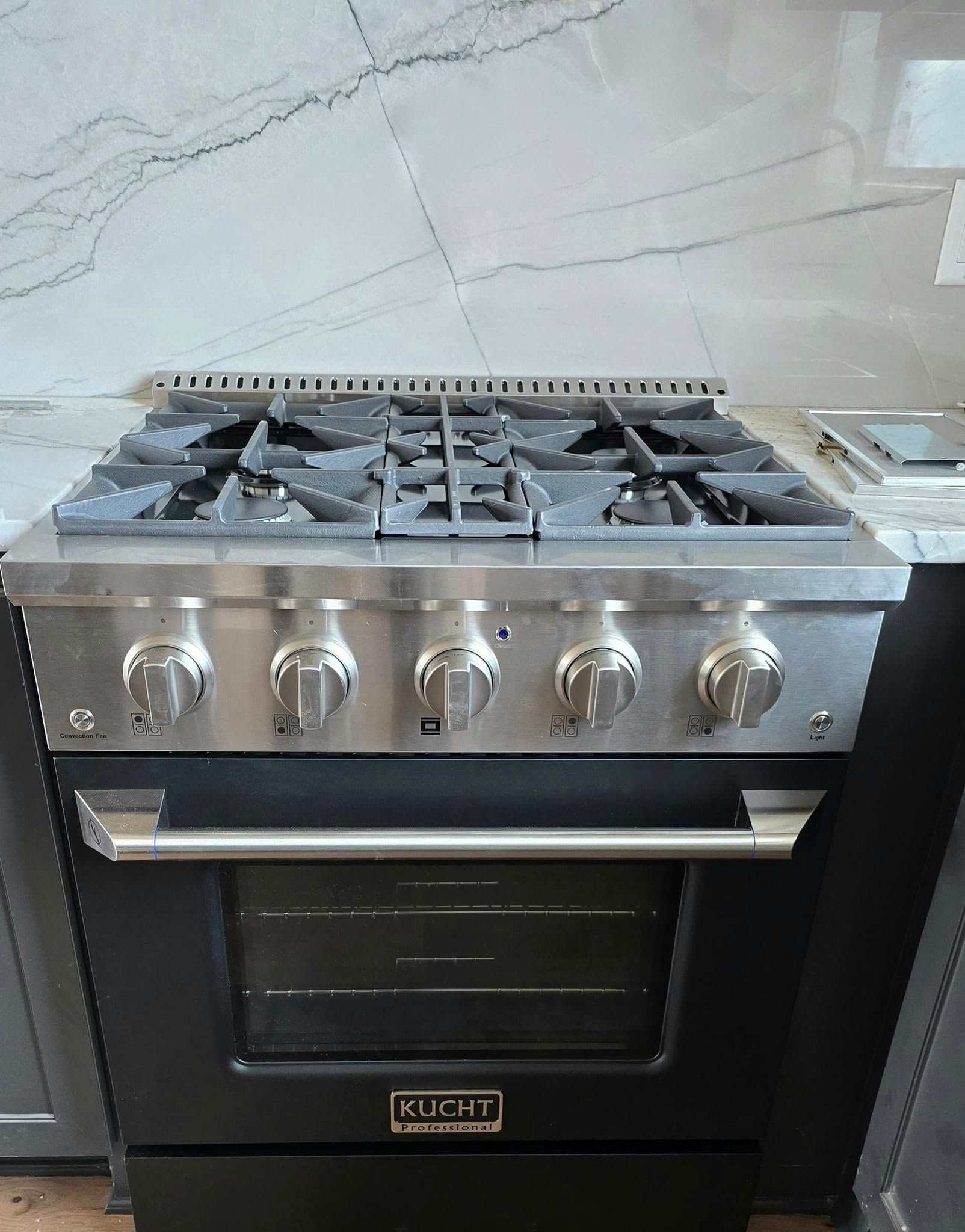 Stainless steel gas range in black cabinet, with five burners and oven. Gray granite countertop.