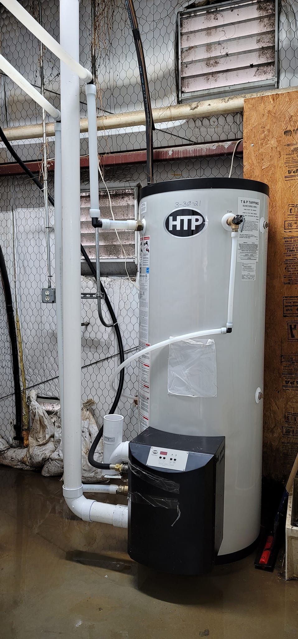 A tall, white HTP water heater in a basement. White pipes are connected to the heater.