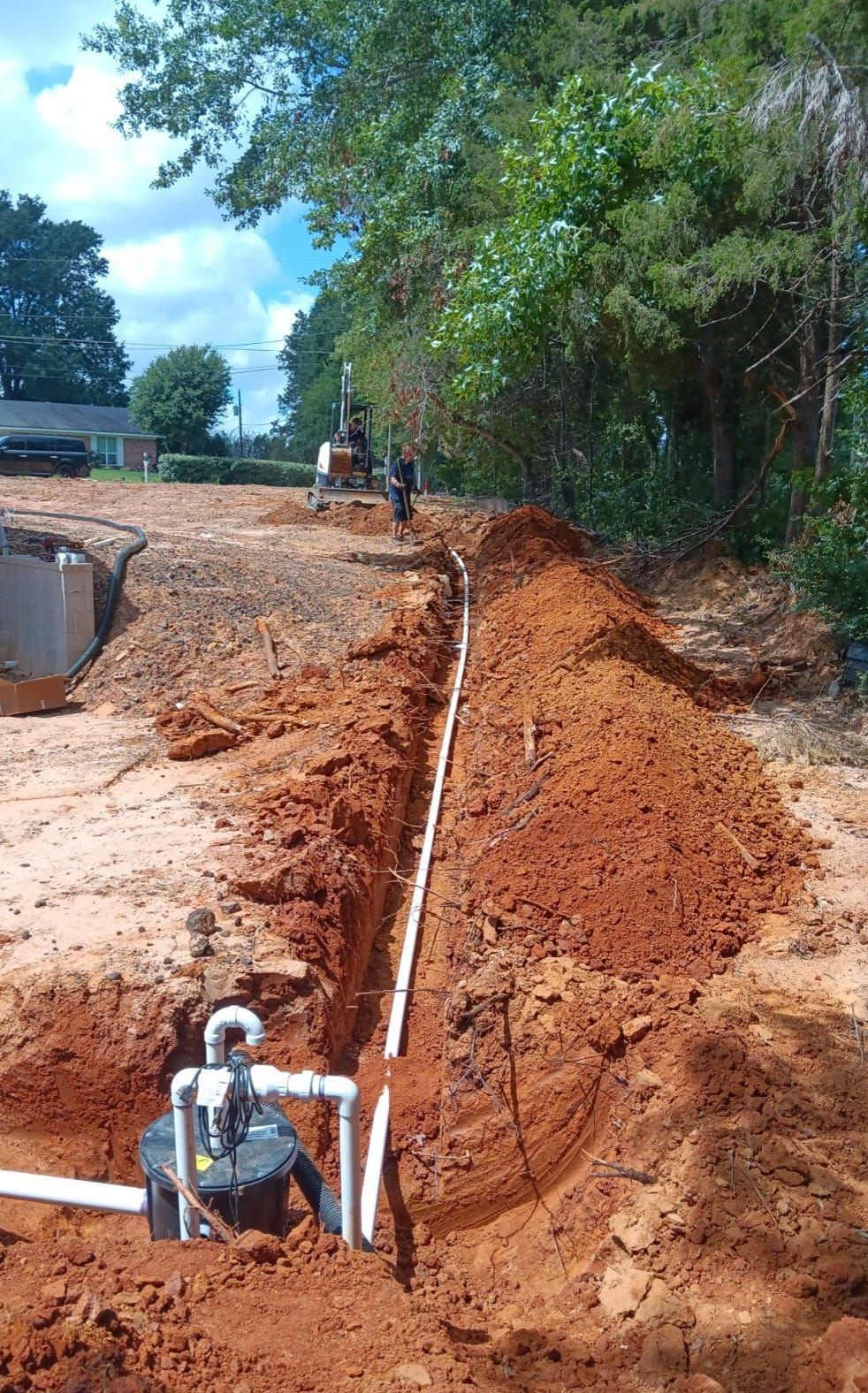 Trench dug in red clay soil with white pipes running through it, a pump is visible in the foreground.