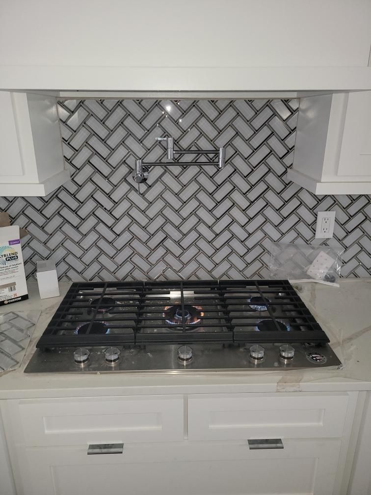 White kitchen with herringbone tile backsplash, gas cooktop, and pot filler faucet.
