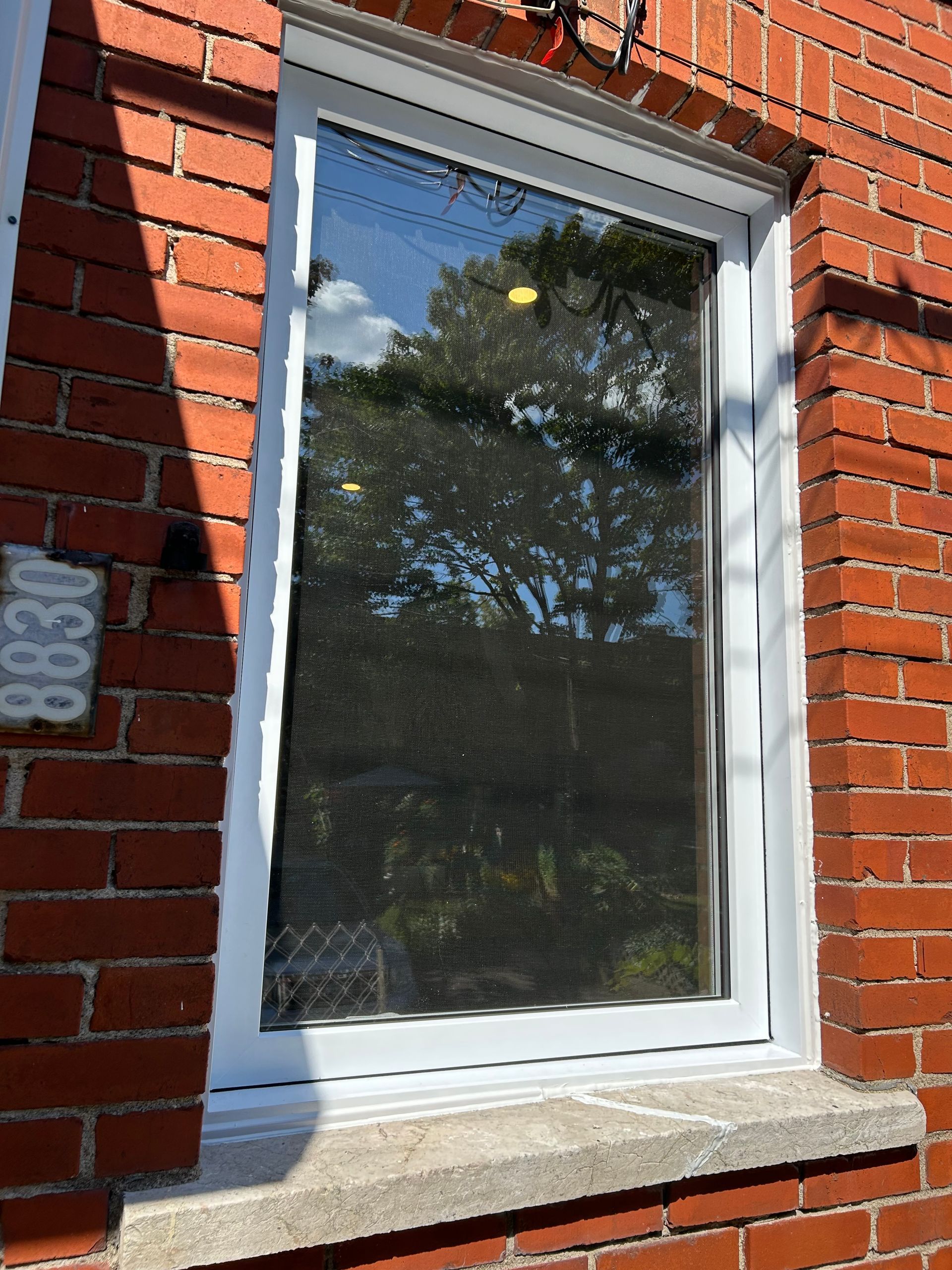 Fenêtre à cadre blanc dans un immeuble en briques. Reflets des arbres, du rebord en béton et du numéro de la maison 8830.