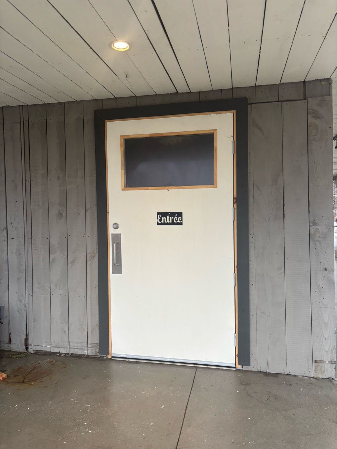 Porte blanche avec une petite fenêtre au-dessus, étiquetée « Entrée » dans un mur lambrissé de bois gris.