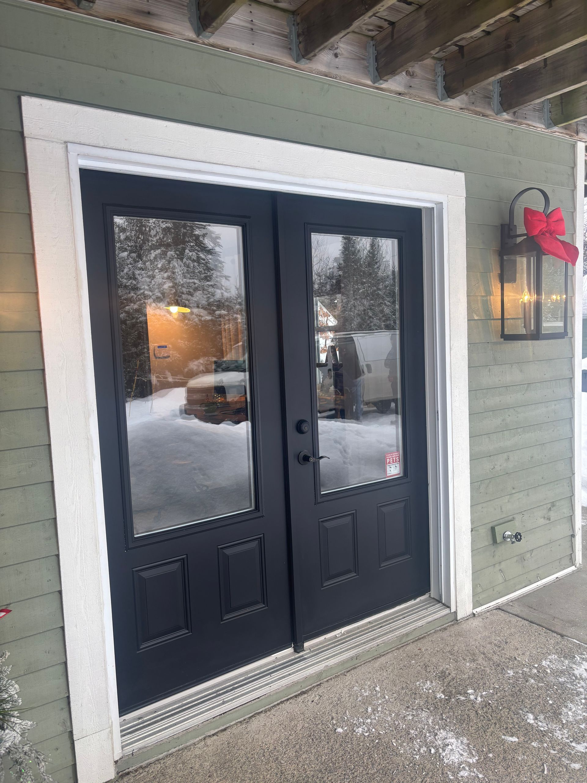 Double porte noire à panneaux de verre et encadrement blanc, montée sur un mur extérieur vert clair ; neige au sol.
