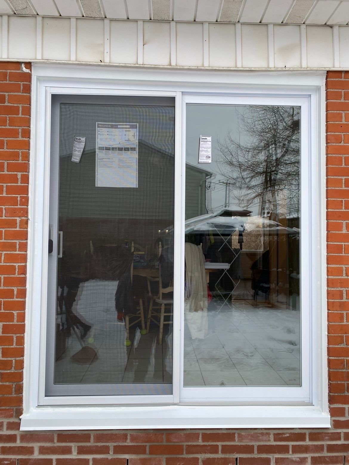 Fenêtre coulissante à cadre blanc dans un mur de briques, un panneau étant muni d'une moustiquaire, reflétant le paysage extérieur et la neige.