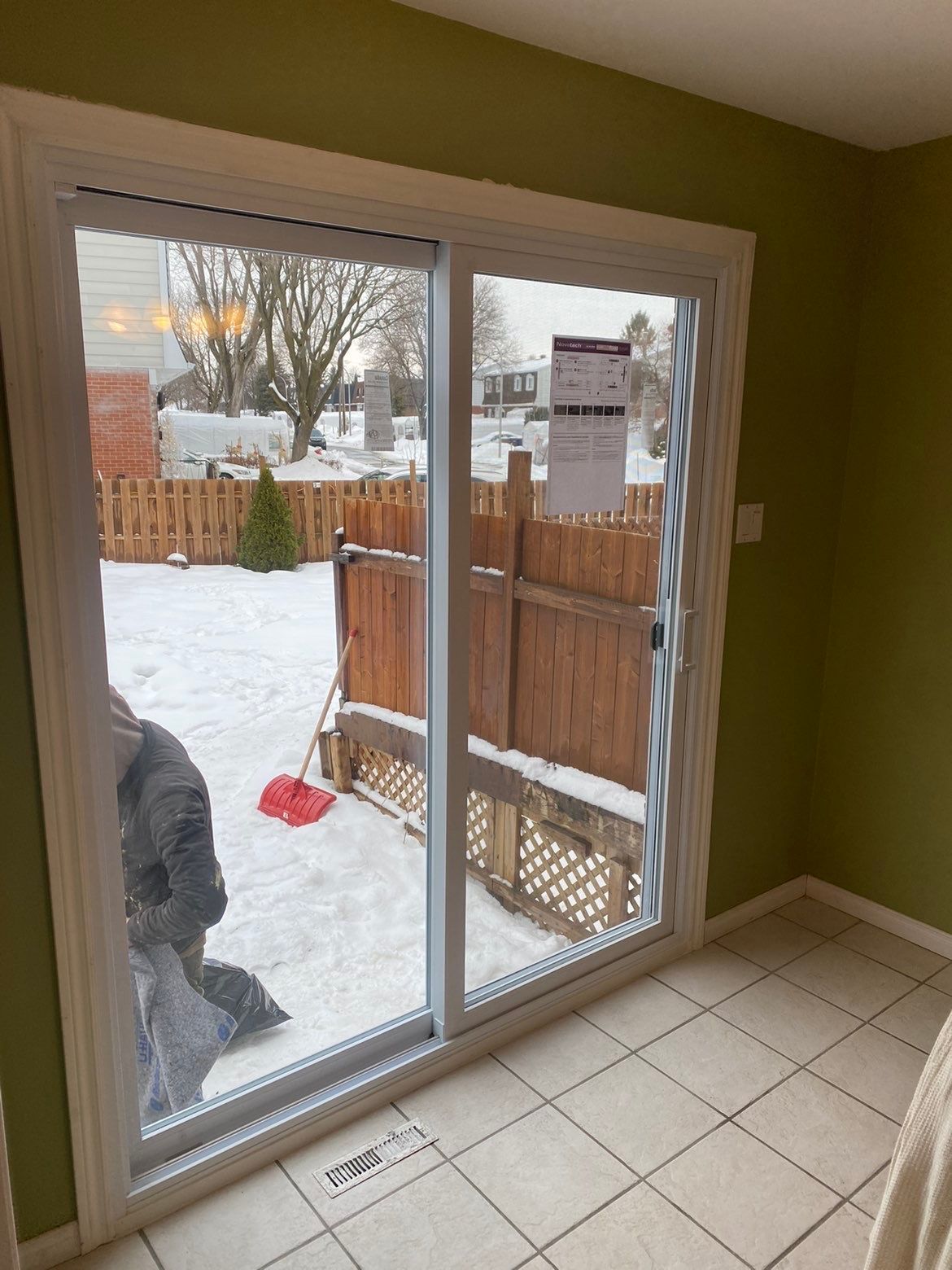Porte-fenêtre coulissante donnant sur un jardin enneigé. Une personne déneige. Murs verts, boiseries blanches.