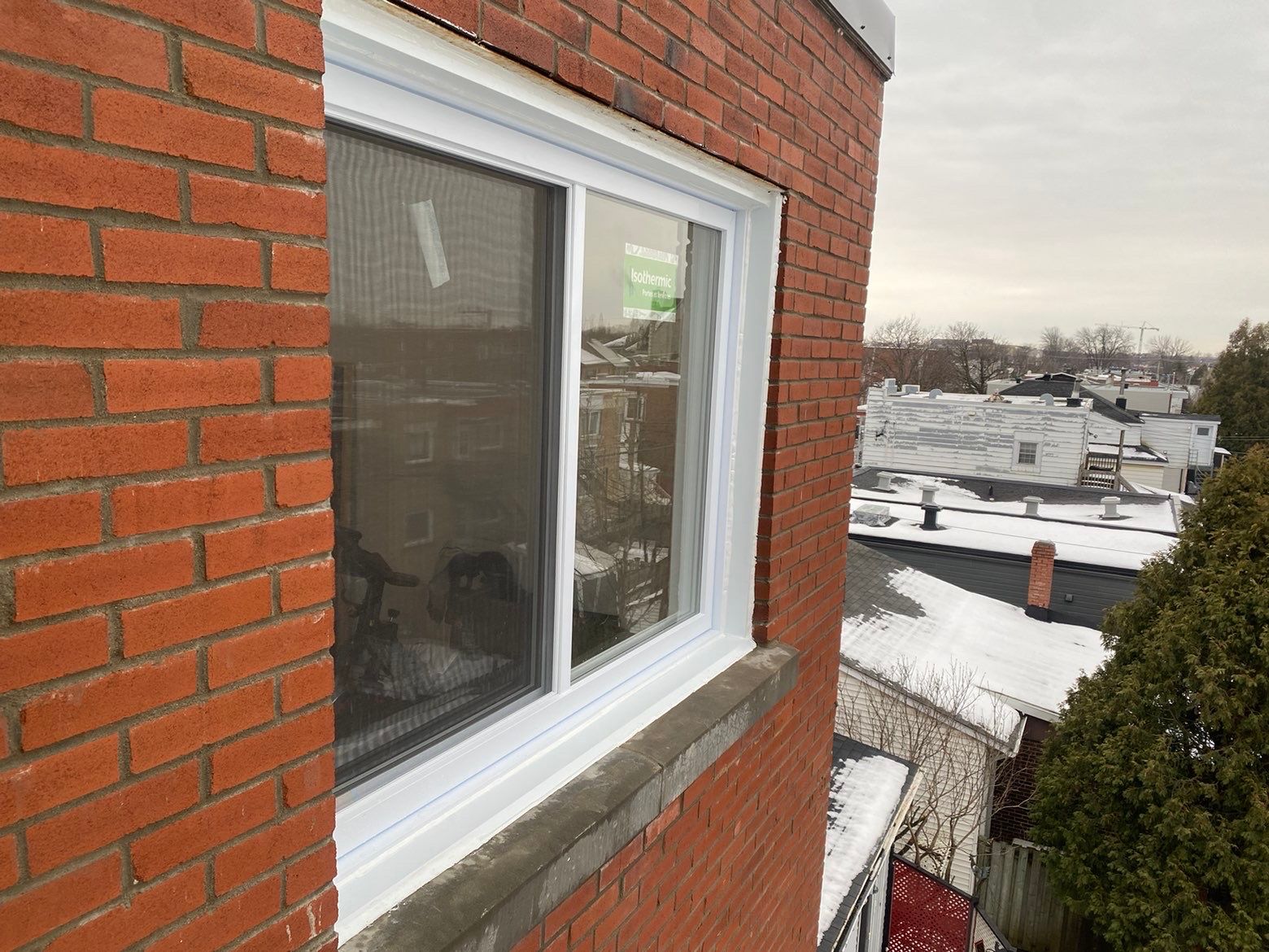 Fenêtre à cadre blanc sur la façade d'un bâtiment en briques rouges, donnant sur des toits enneigés.