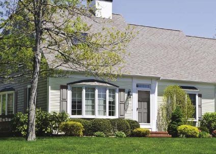 Une maison claire avec un toit gris et des baies vitrées, entourée d'herbe verte et de buissons.