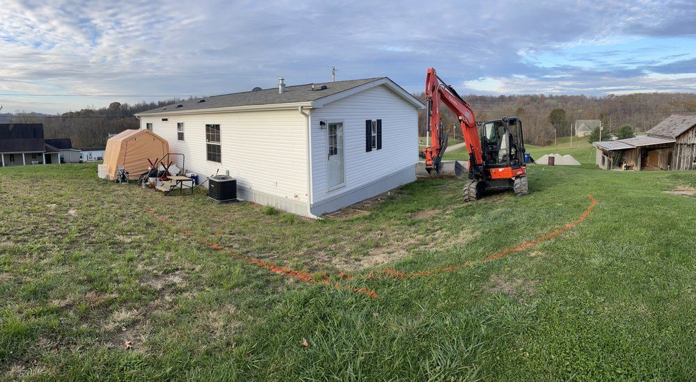 A small excavator is sitting in front of a white house in a grassy field.