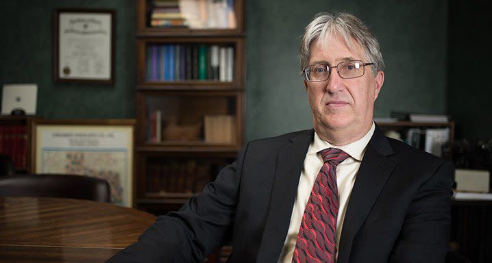 A man in a suit and tie is sitting at a table in front of a bookshelf.