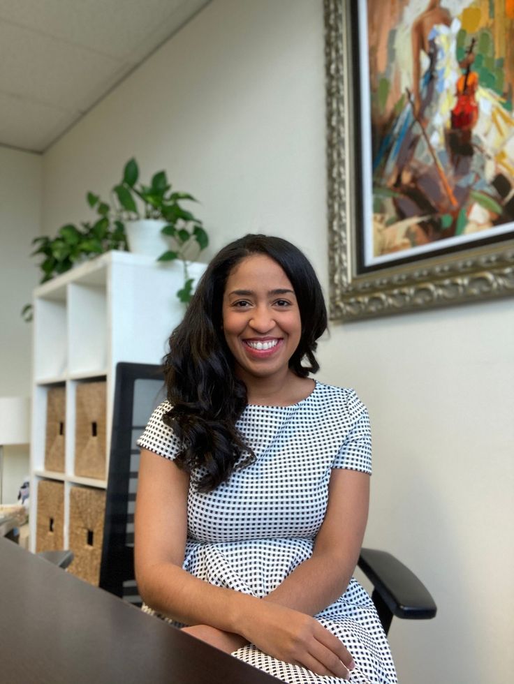 Smiling woman with long black hair, wearing black scrubs, sitting in a blue chair.