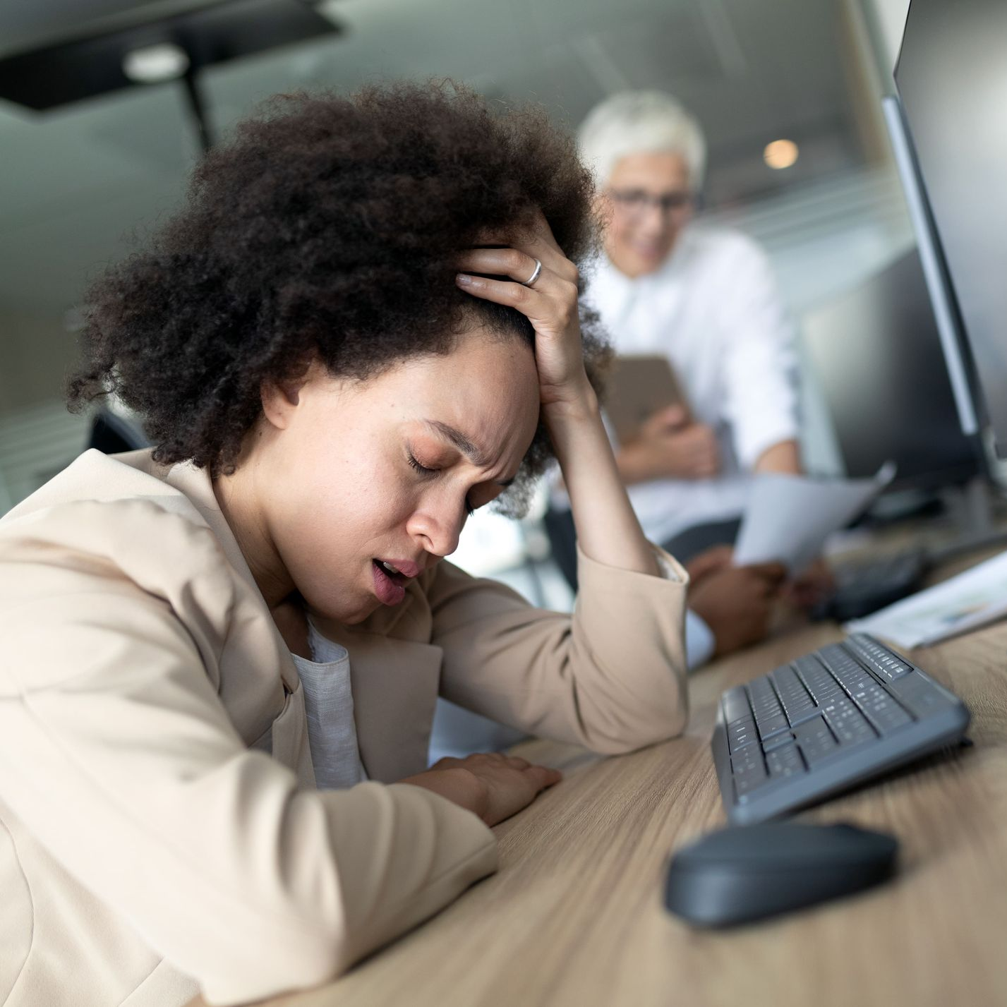 Woman with head in hand, stressed at desk with keyboard; colleague in background.