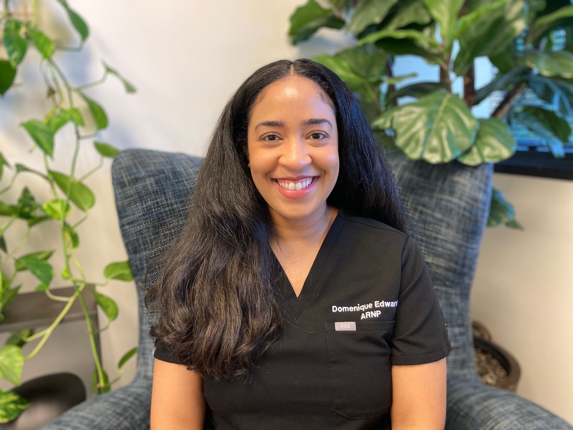 Smiling woman with long black hair, wearing black scrubs, sitting in a blue chair.