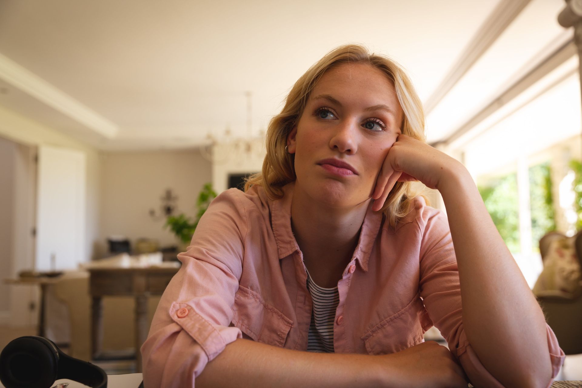 Blonde woman resting head on hand, looking bored. Wearing pink shirt, sitting indoors.