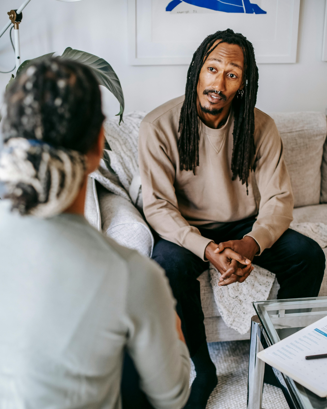 Man with dreadlocks and woman converse on a couch; indoor setting.