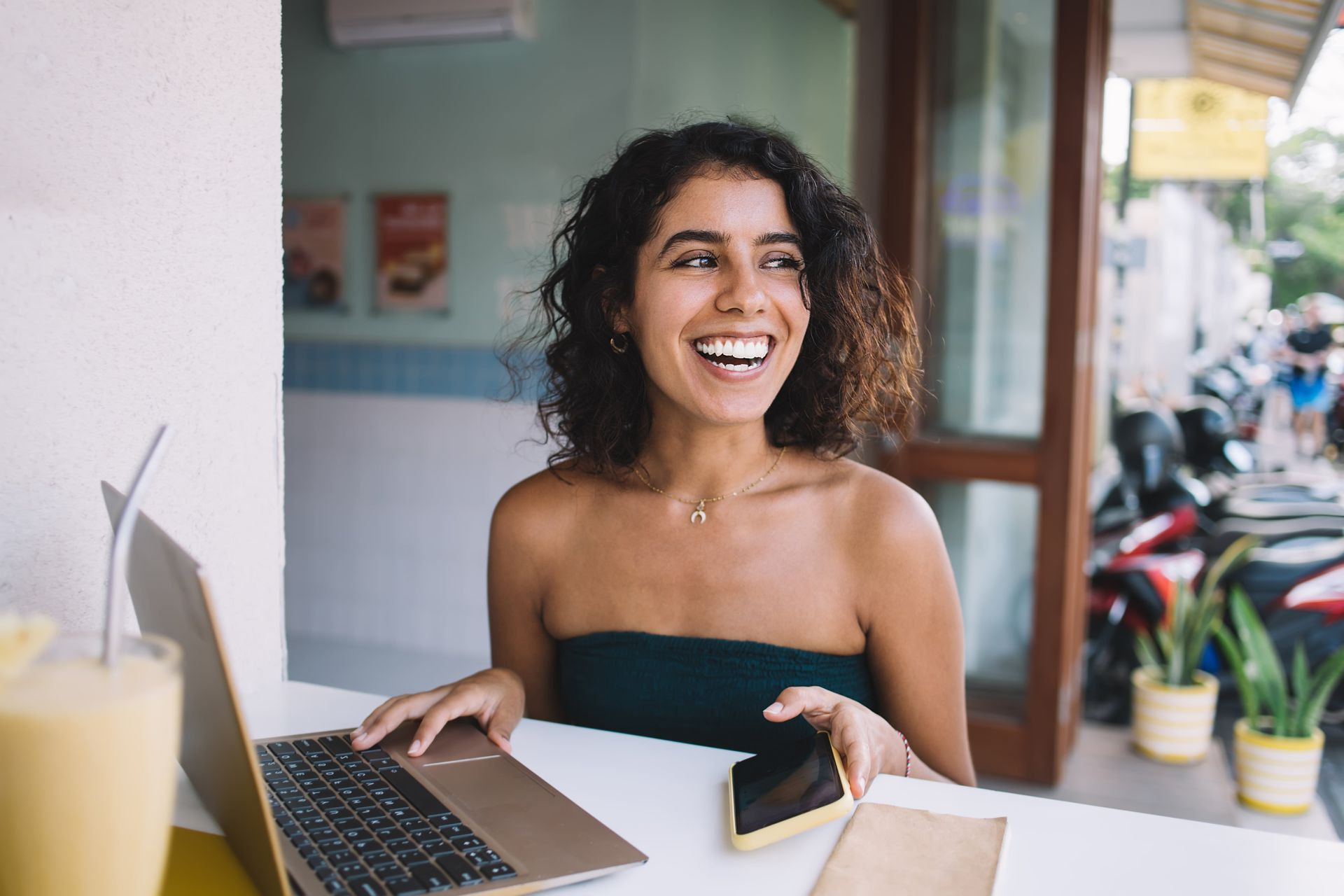Smiling person at a café table with a laptop and coffee, looking toward the street.