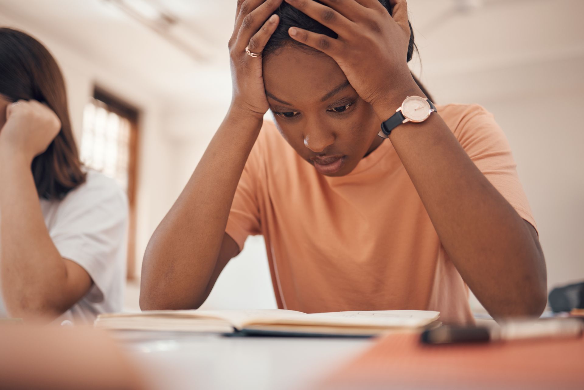 Woman stressed over book, hands on head, indoors with another person, orange shirt, watch.