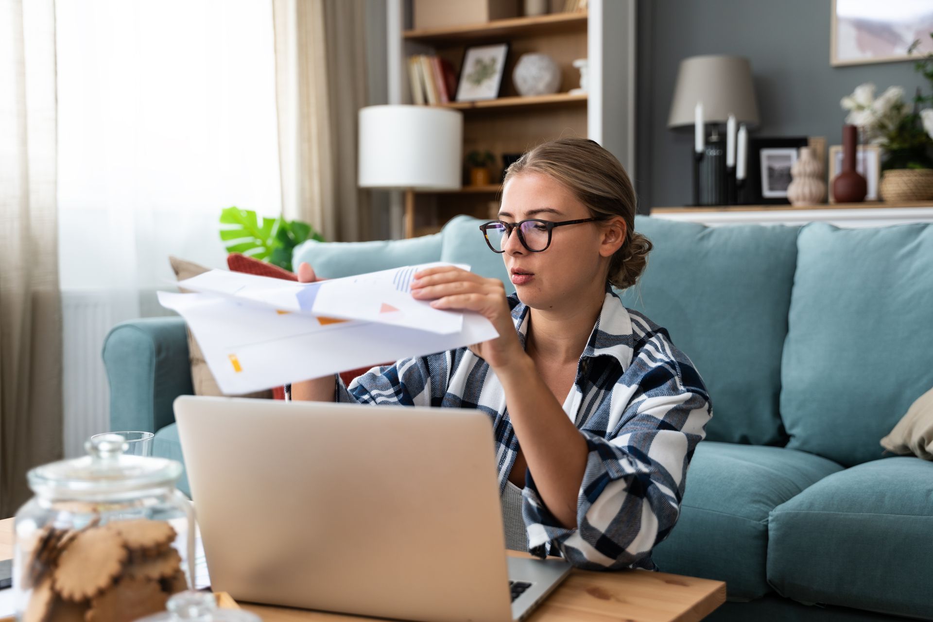 Woman in glasses reviewing papers, sitting on floor by laptop in living room.