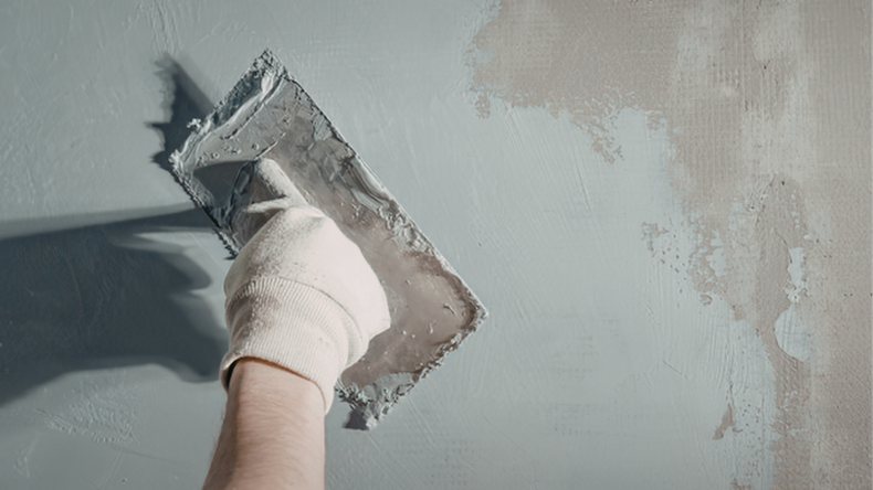 Gloved hand smoothing gray plaster on a wall with a trowel