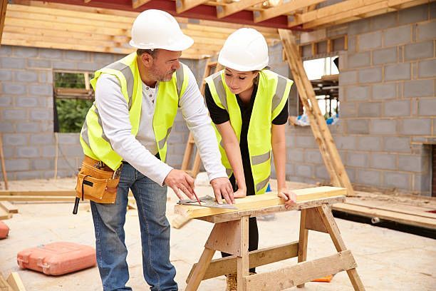 Two construction workers in hard hats measuring wood on a sawhorse inside a building under construction