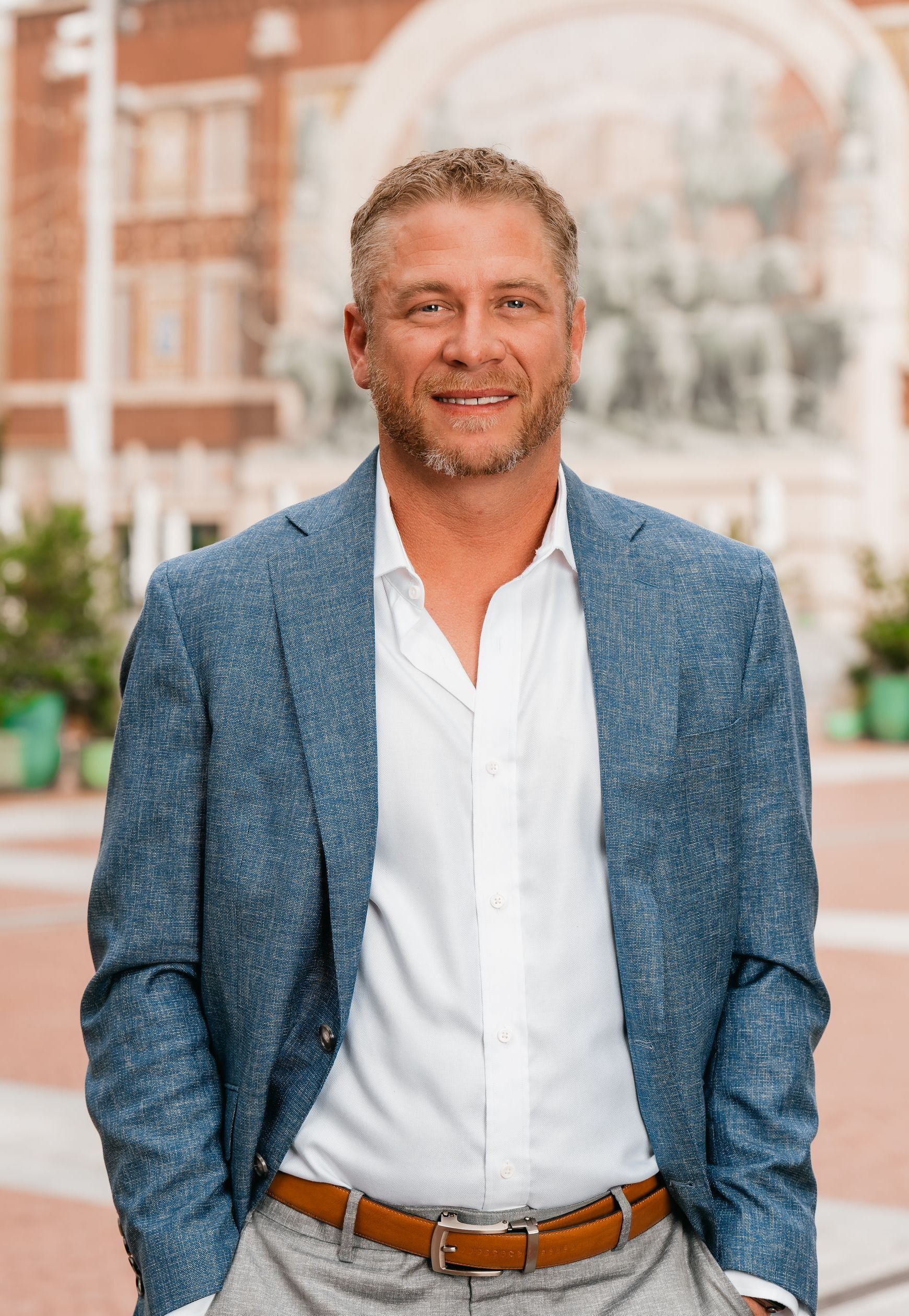 A man in a blue jacket and white shirt is standing in front of a building.