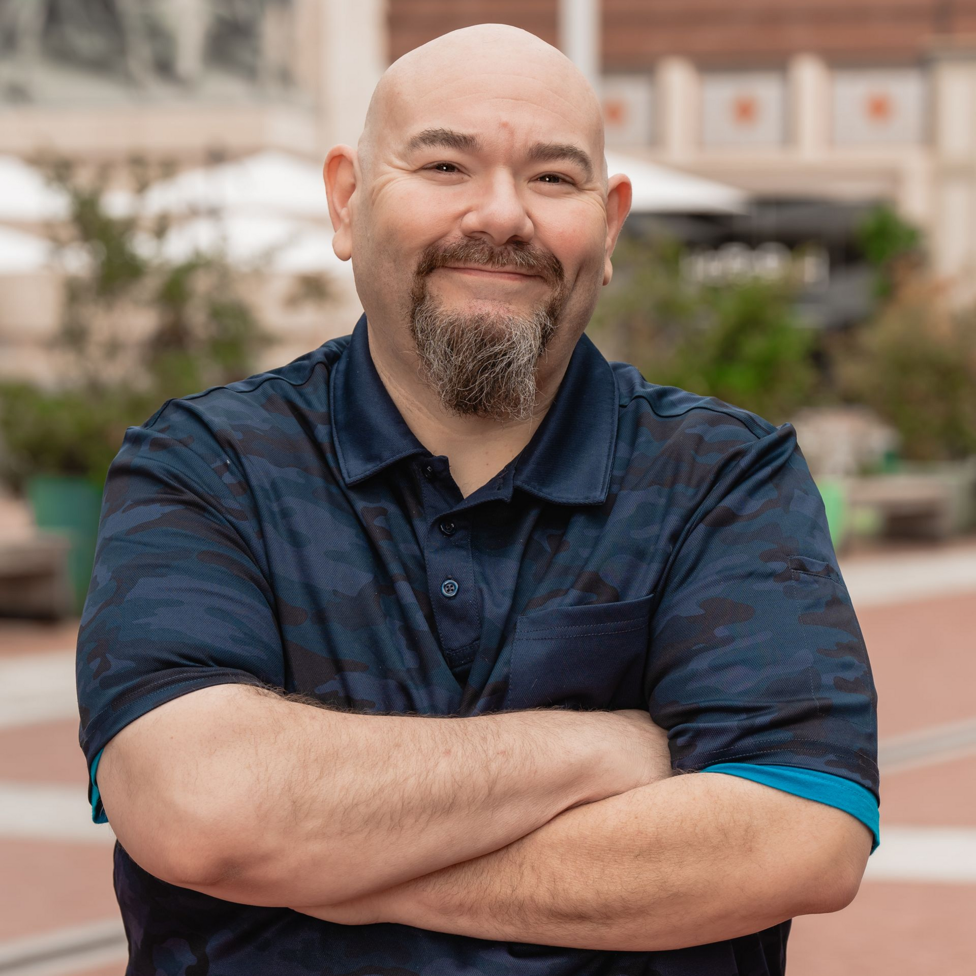 A bald man with a beard is standing with his arms crossed and smiling.