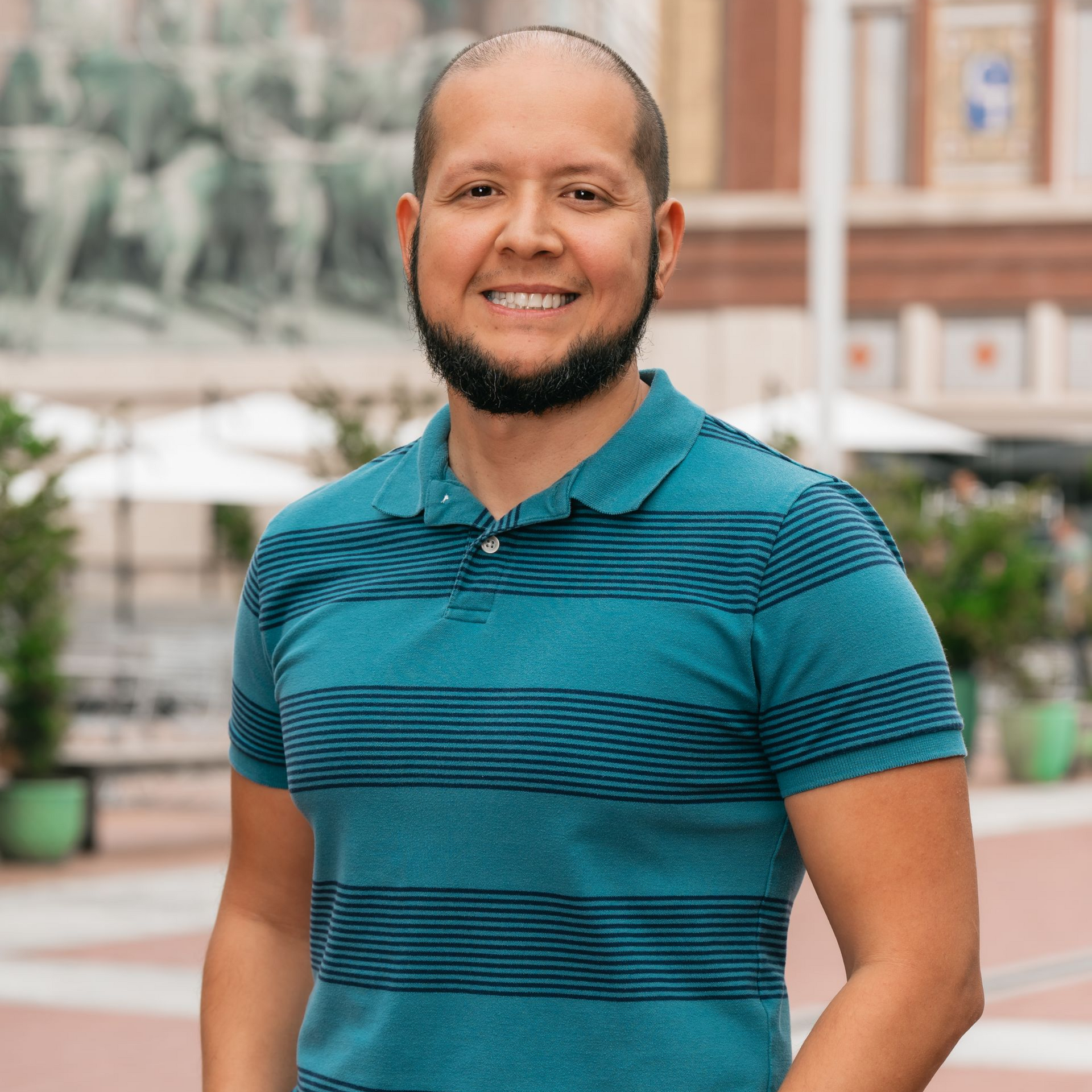 A man with a beard is wearing a blue striped shirt and standing in front of a building.