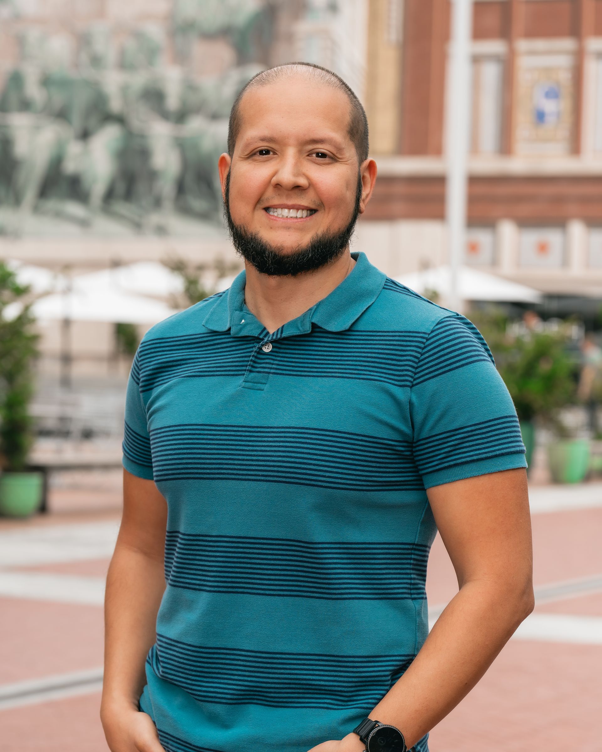 A man with a beard is wearing a blue striped shirt and standing in front of a building.