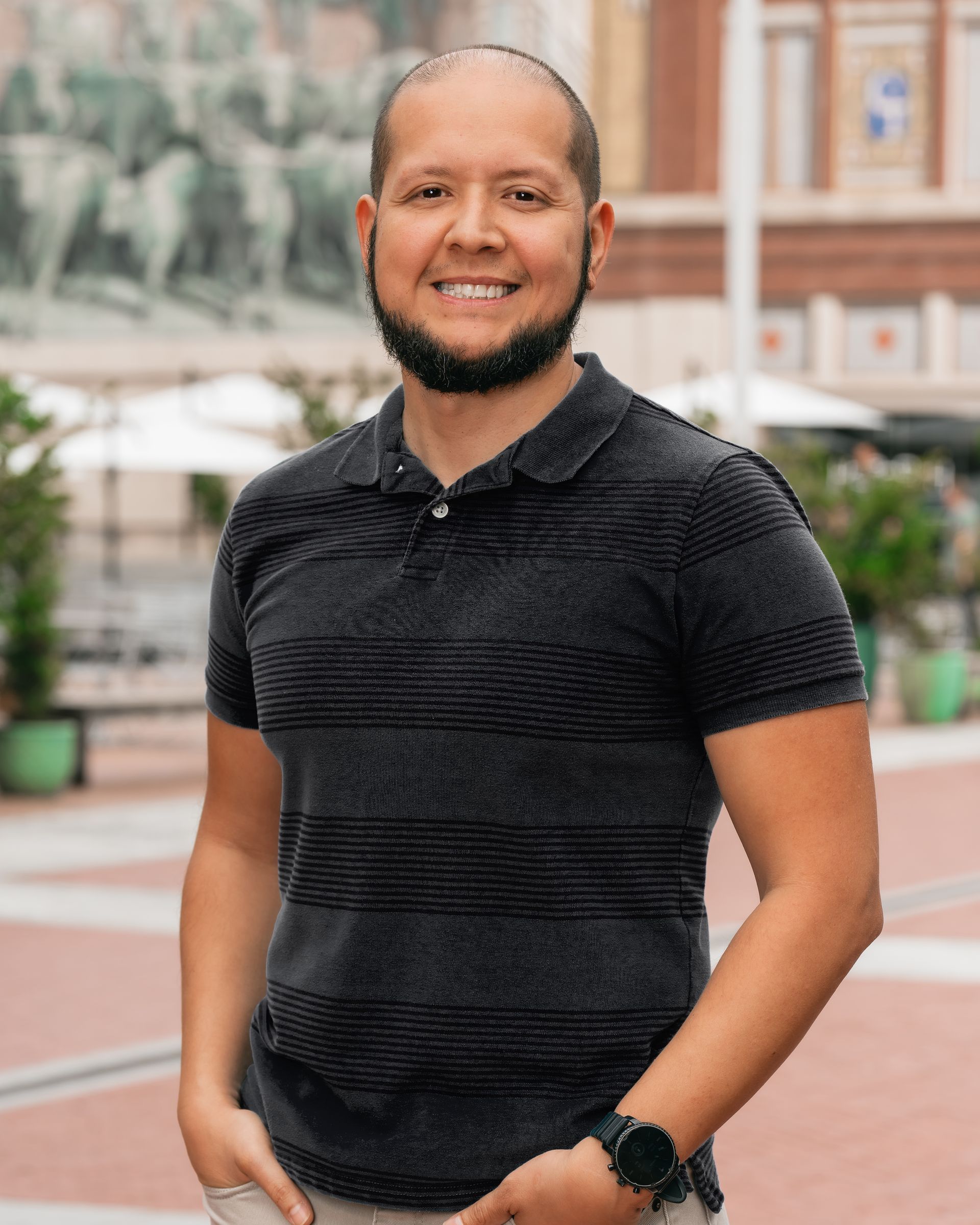 A man with a beard is wearing a blue striped shirt and standing in front of a building.