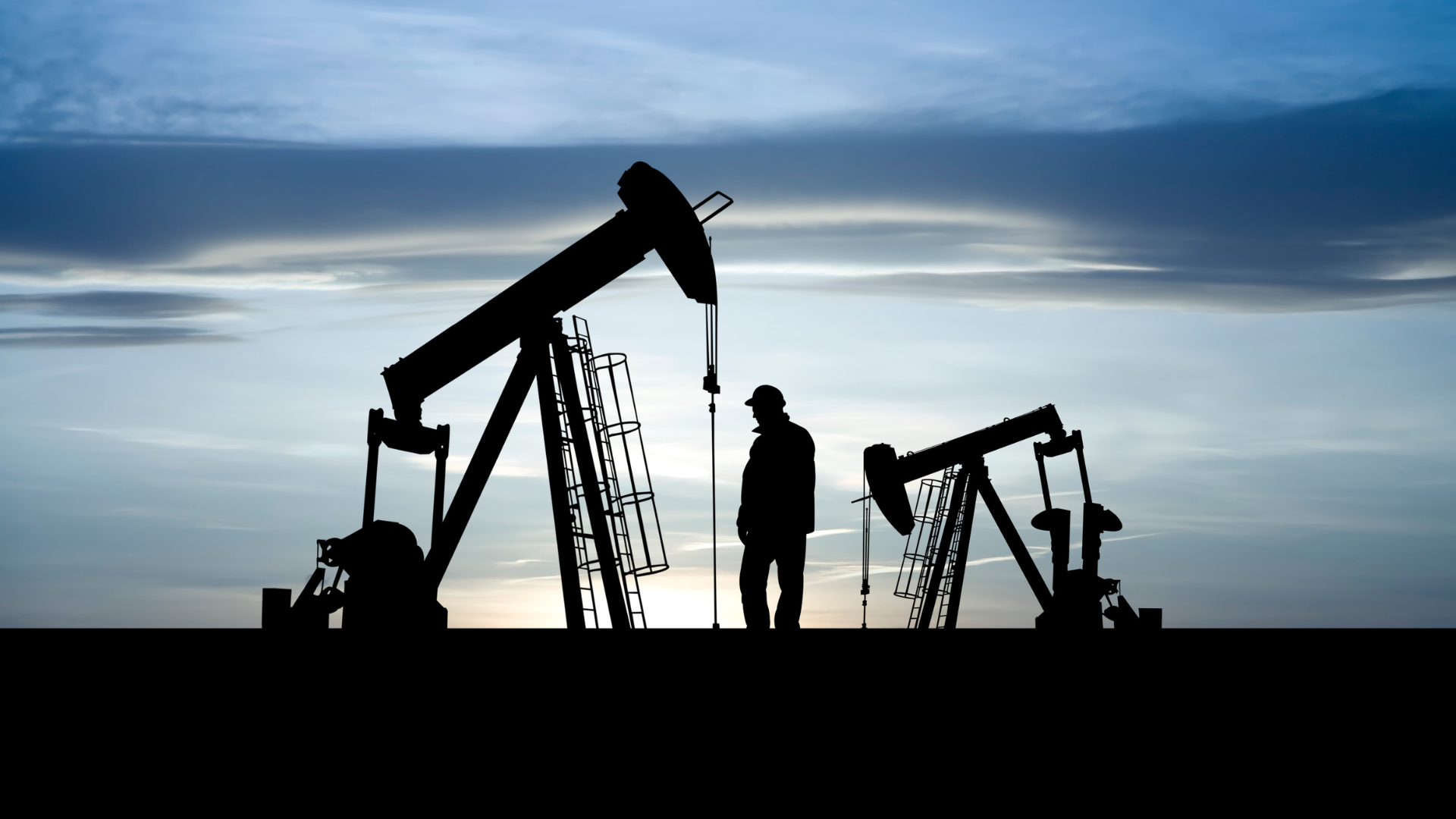 Silhouetted oil pumpjacks with a person standing between them against a dusk sky.