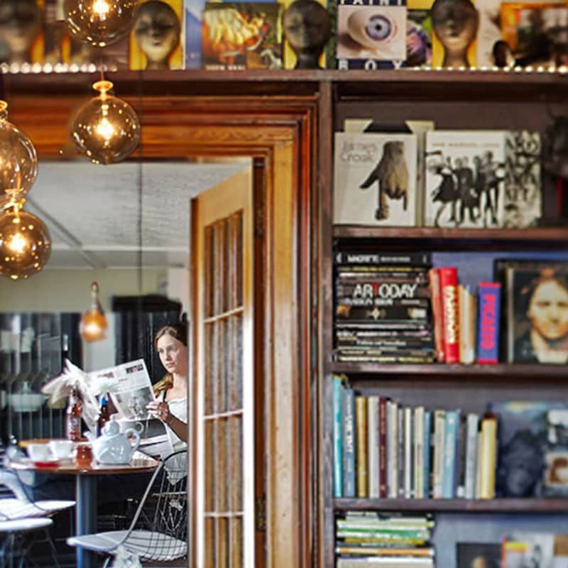 A woman sitting at a table reading a newspaper next to a bookshelf that has ar today on it