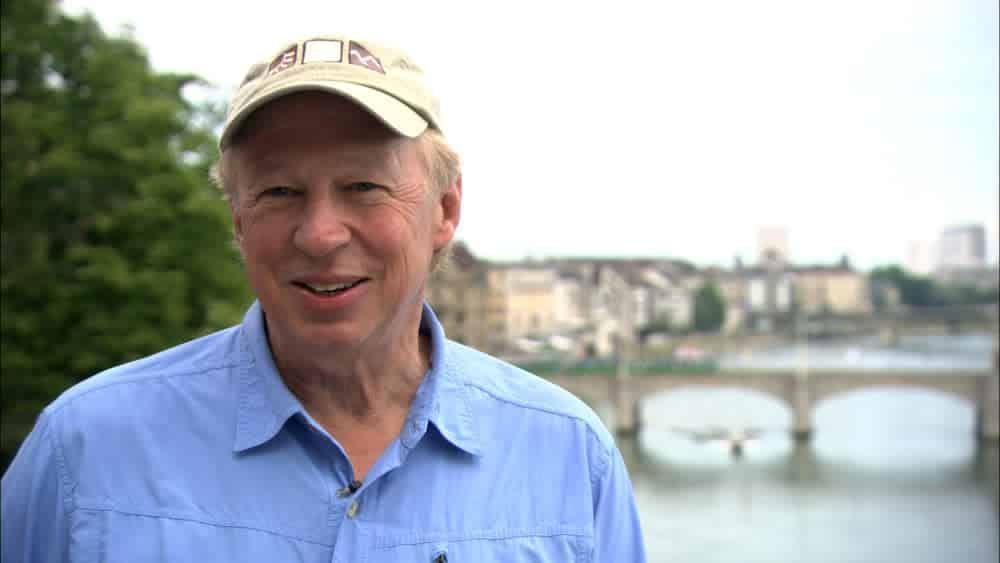A man wearing a hat and a blue shirt is standing in front of a river.