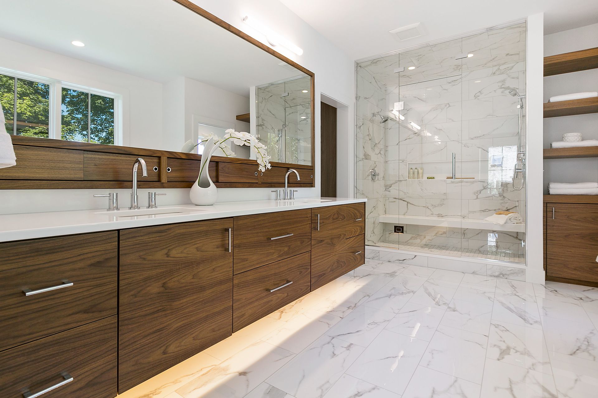 Modern bathroom with wooden vanity, large mirror, marble shower, and flooring.