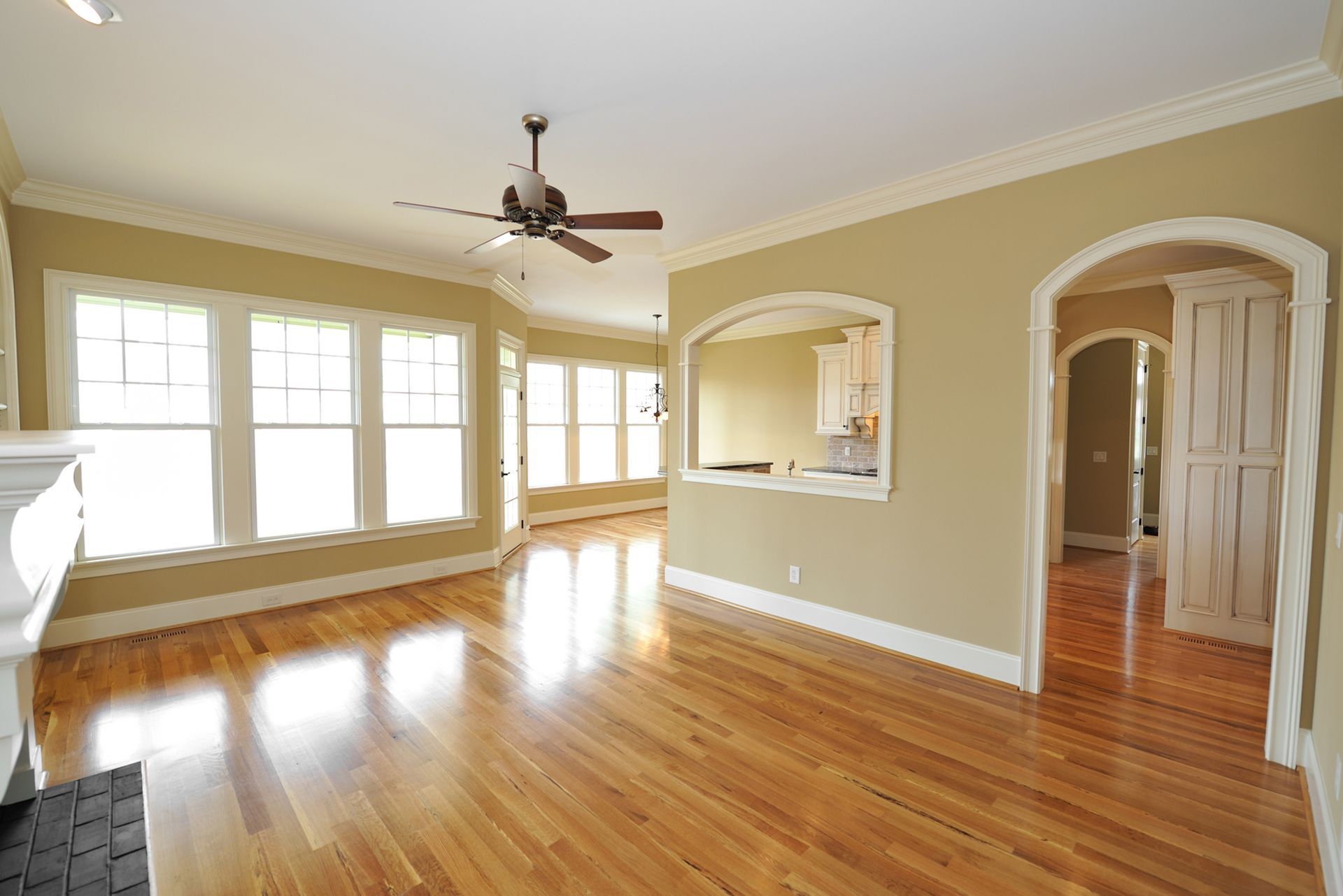 Spacious, sunlit room with wood floors, large windows, beige walls, and arched doorways.