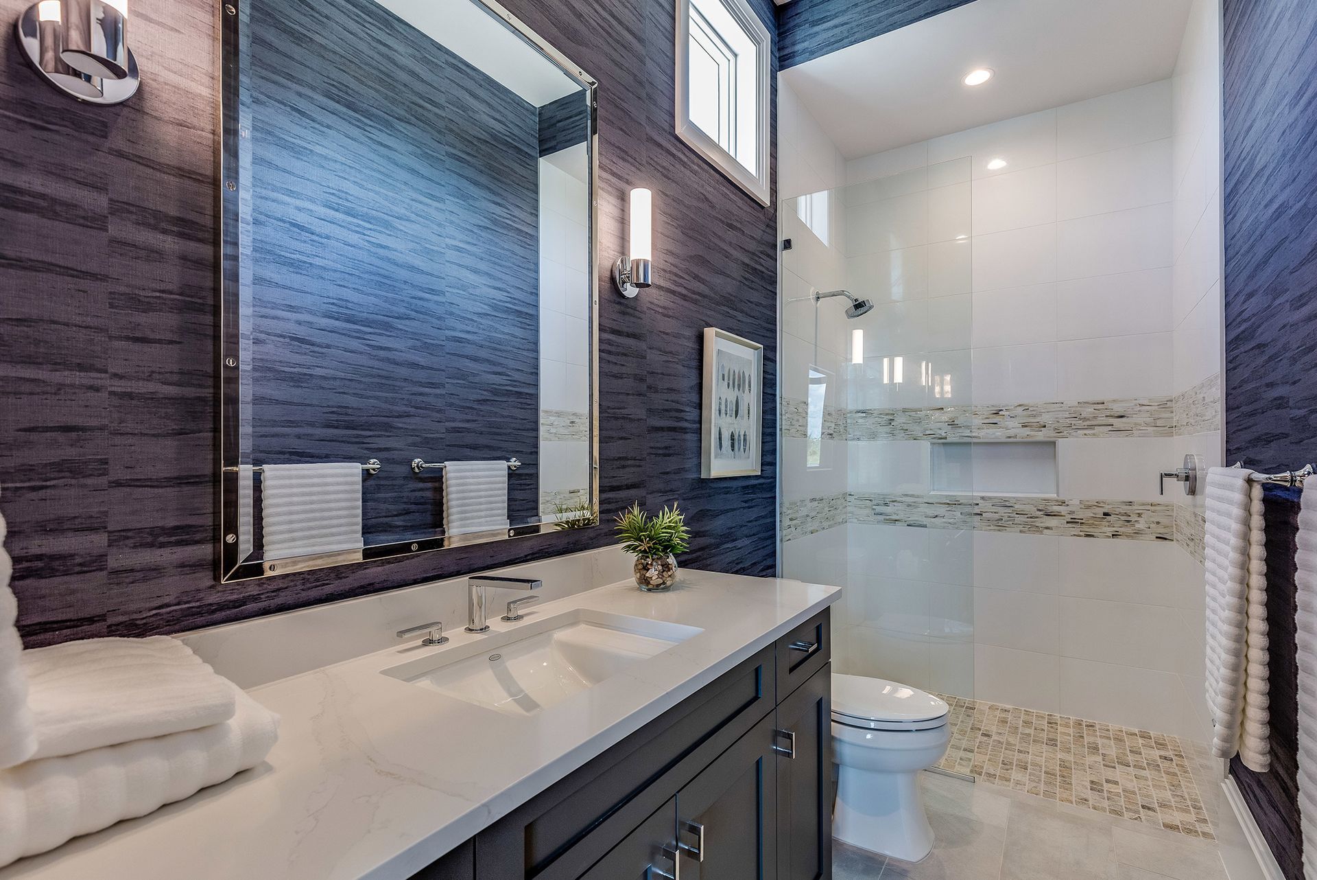 Modern bathroom with navy blue textured walls, white countertops, glass shower, and pebble floor.