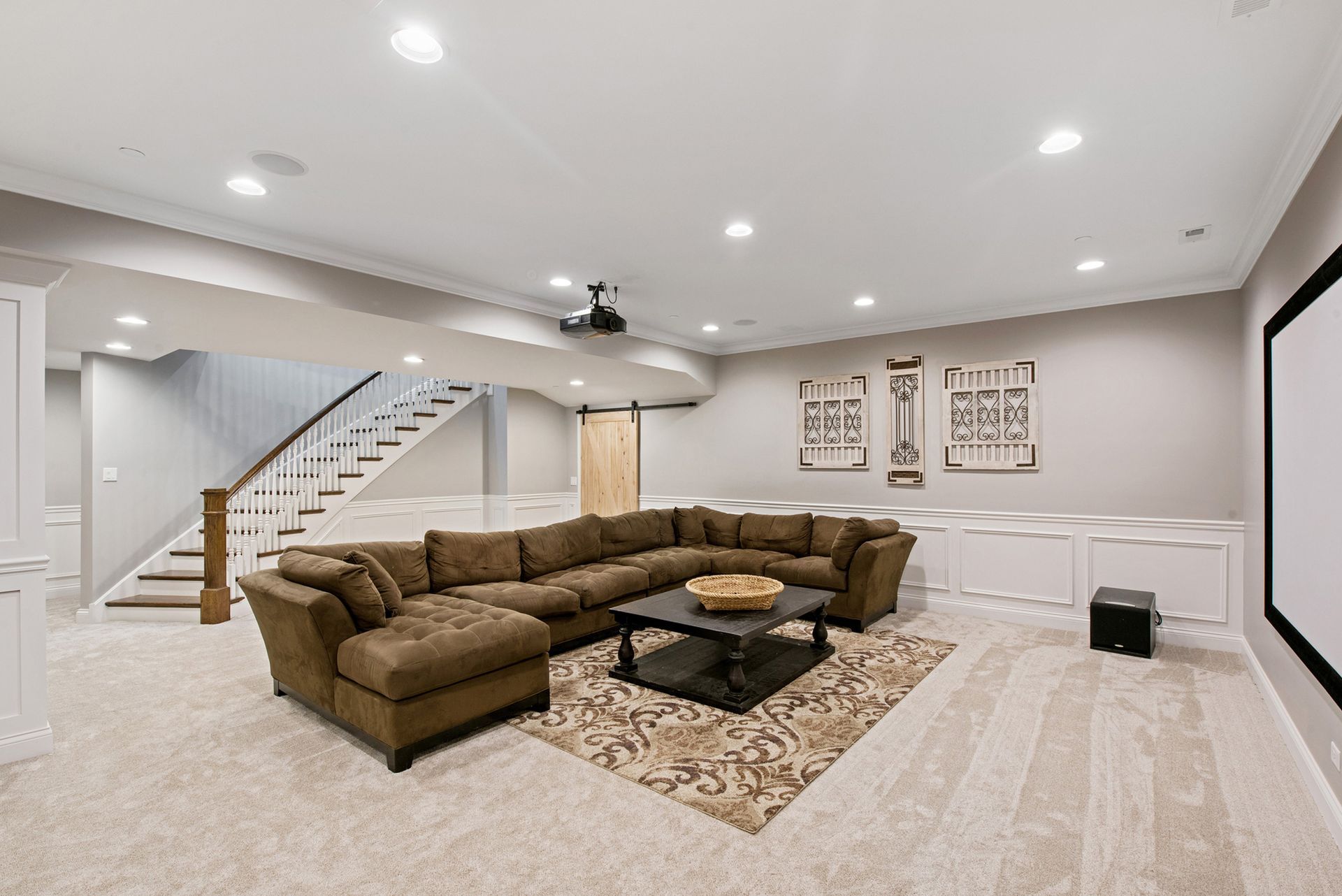 A cozy basement entertainment room with a sectional sofa, projector, and stairs.