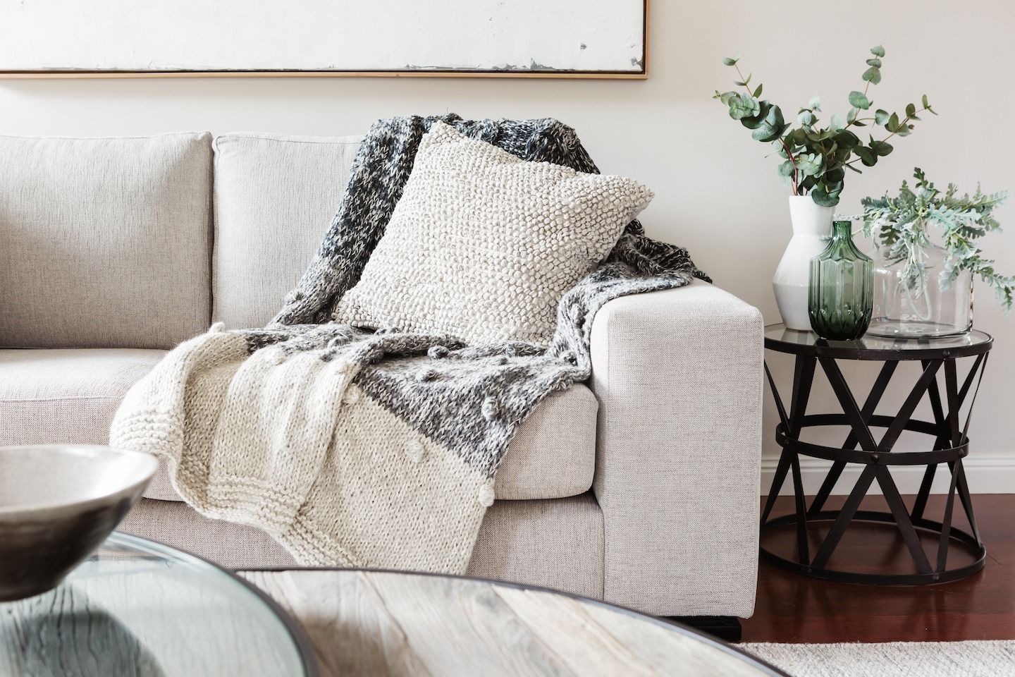 Cozy beige sofa with knit throw, cushion, and side table with vases and greenery.