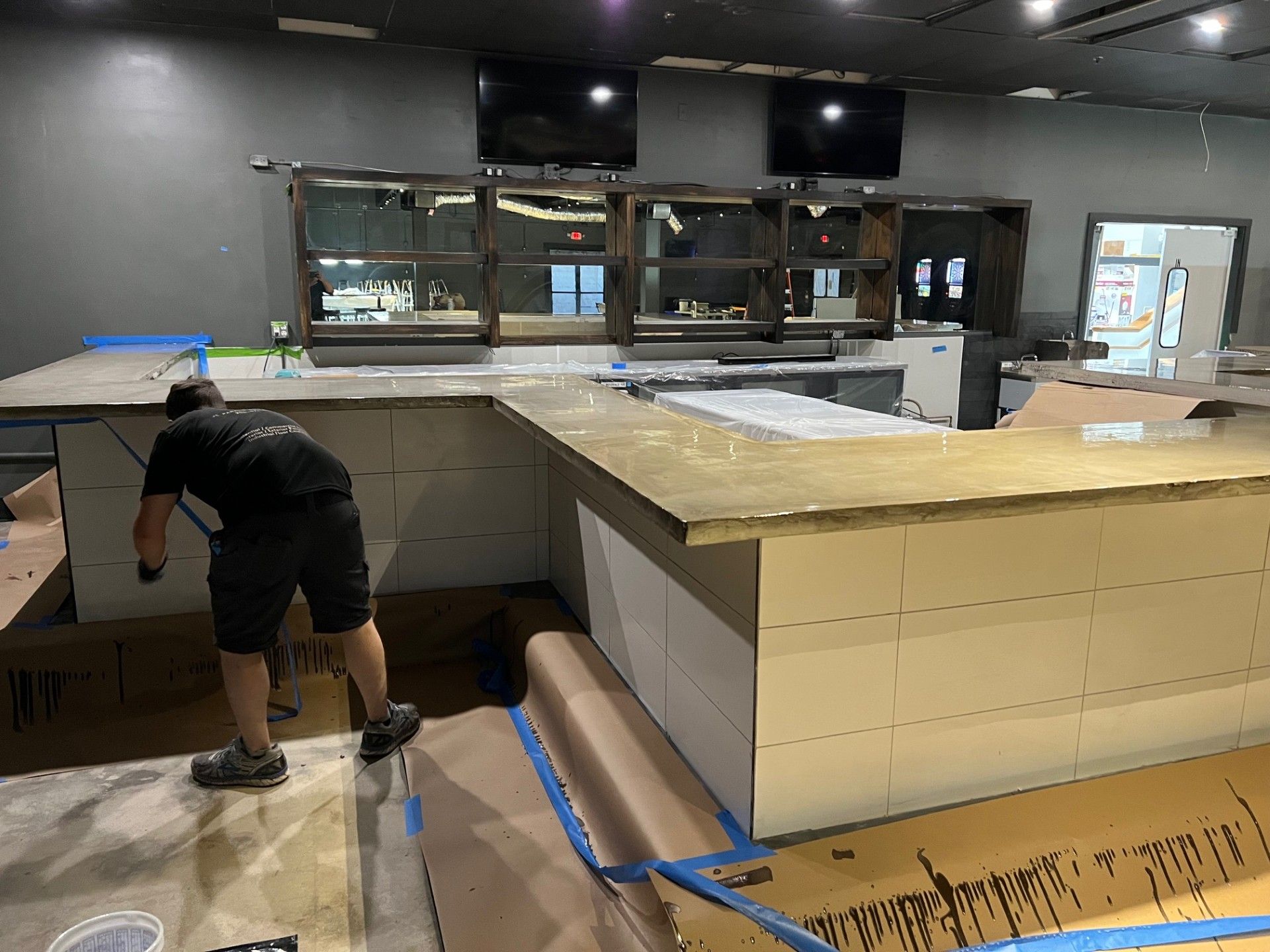 Person installing counter in a bar with dark walls, mirrors, and TVs; unfinished.