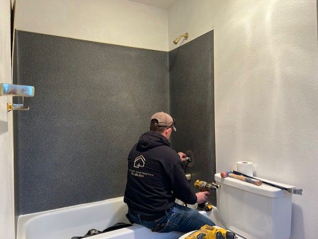 A man in a black hoodie installing shower fixtures in a bathroom with dark gray walls.