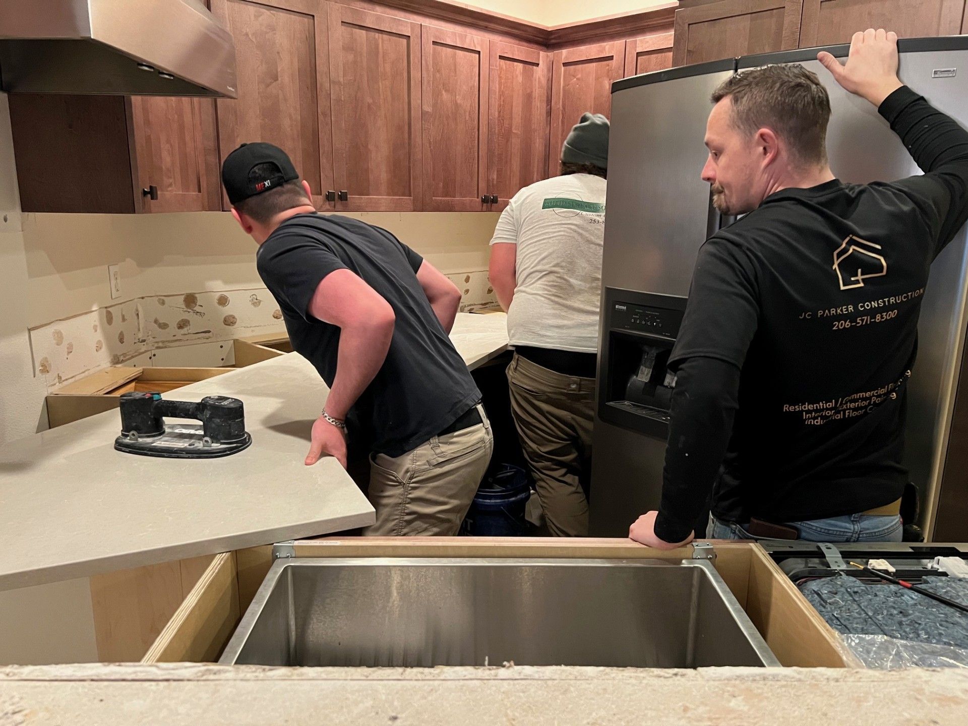 Three workers installing a kitchen countertop and refrigerator; stainless steel sink.