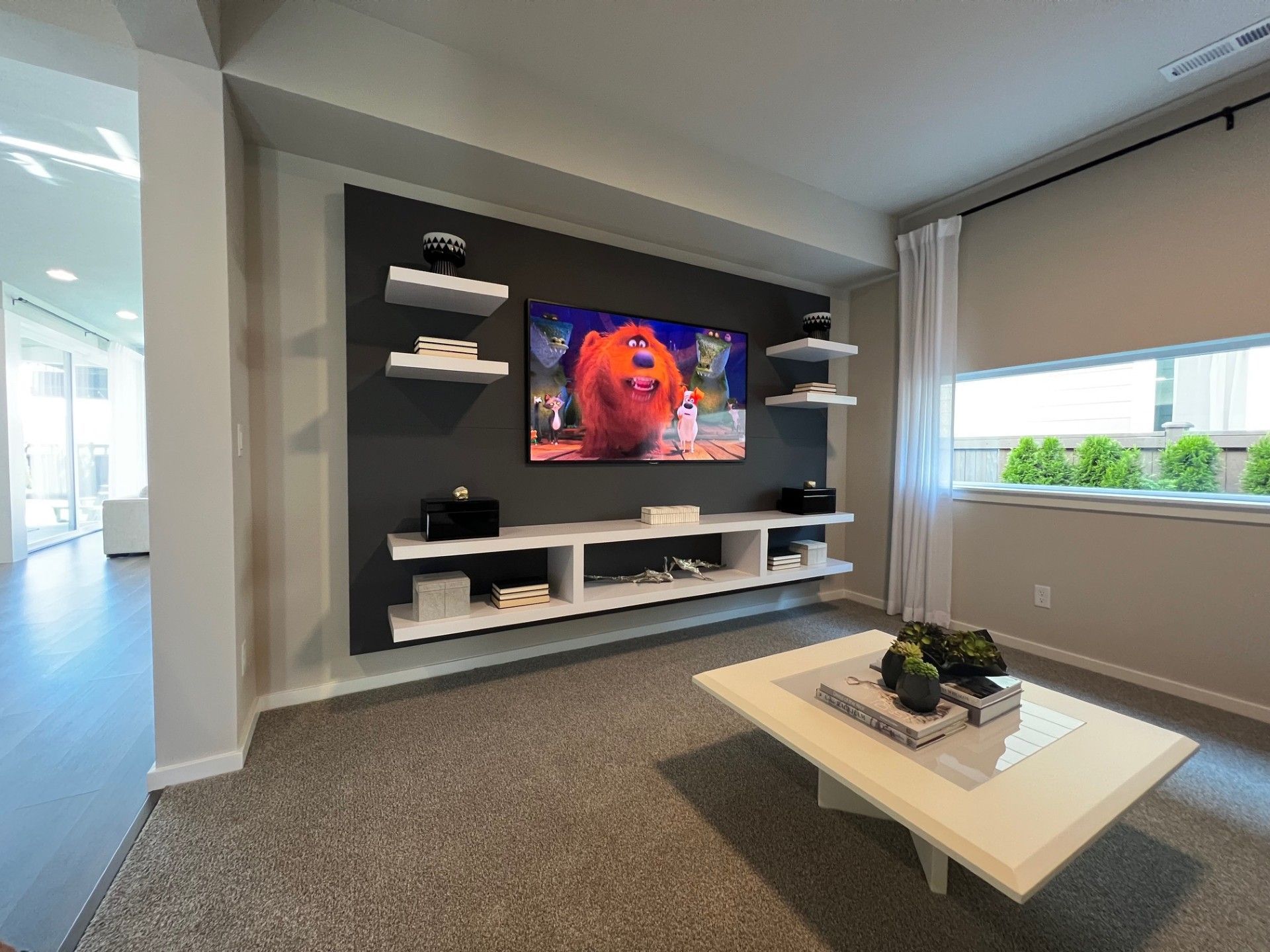 Living room with TV on dark gray wall, white shelves, gray carpet, white table, and movie playing.