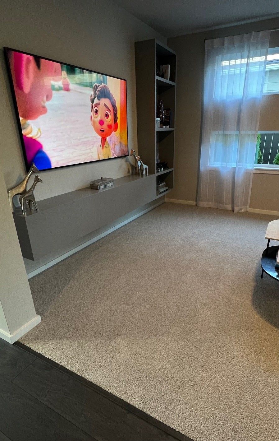 Living room with a large TV on a floating shelf, a bookshelf, and a window with sheer curtains.