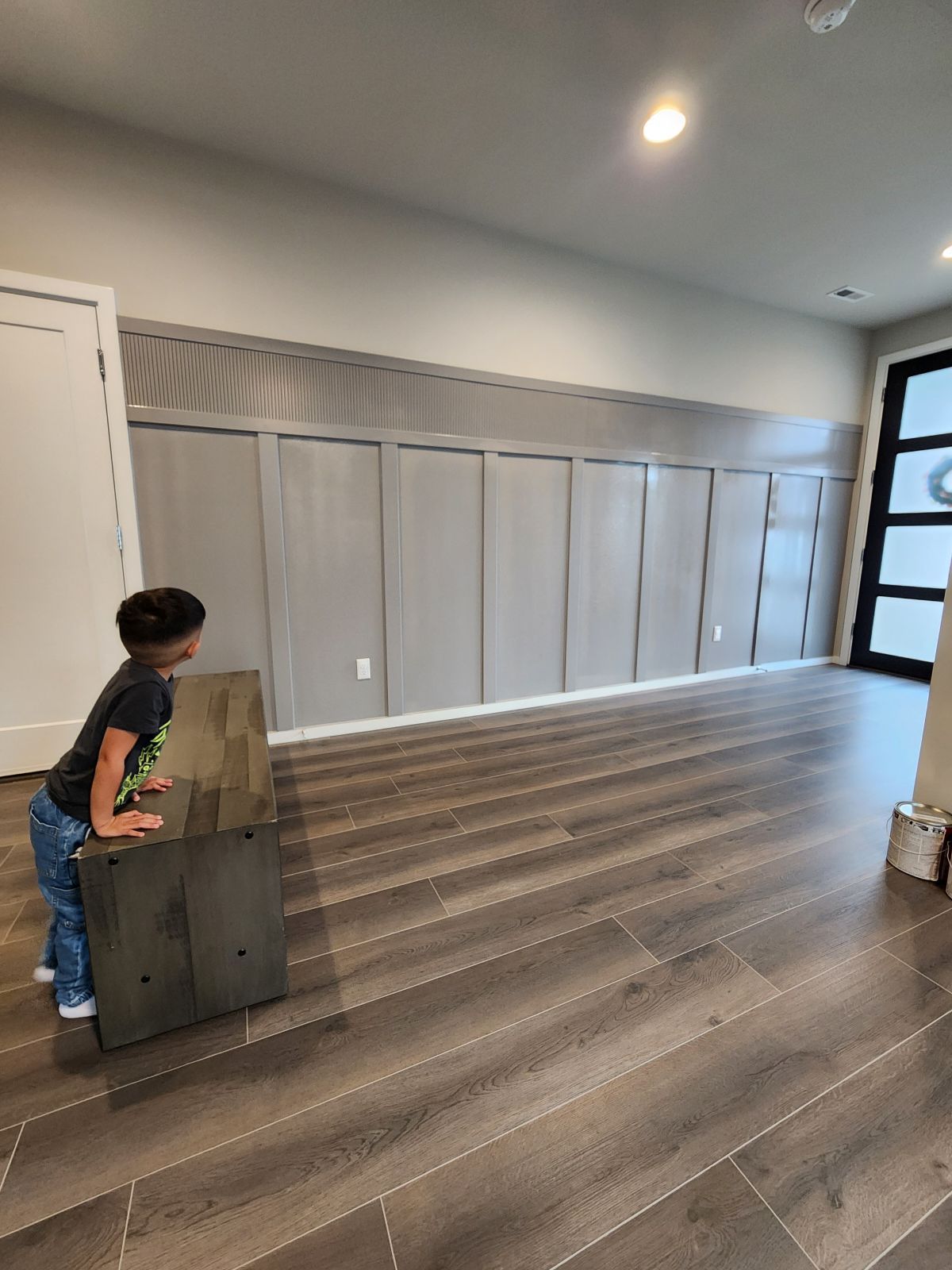 Boy looking at wooden block; gray paneled wall, dark wood floor.