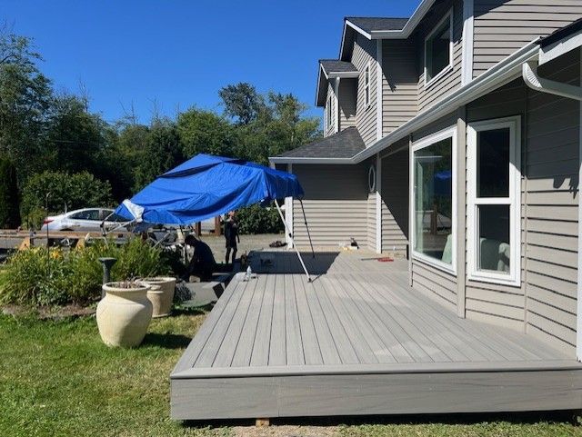 Wooden deck extends from a two-story house; a blue tarp provides shade. People are working on the deck.