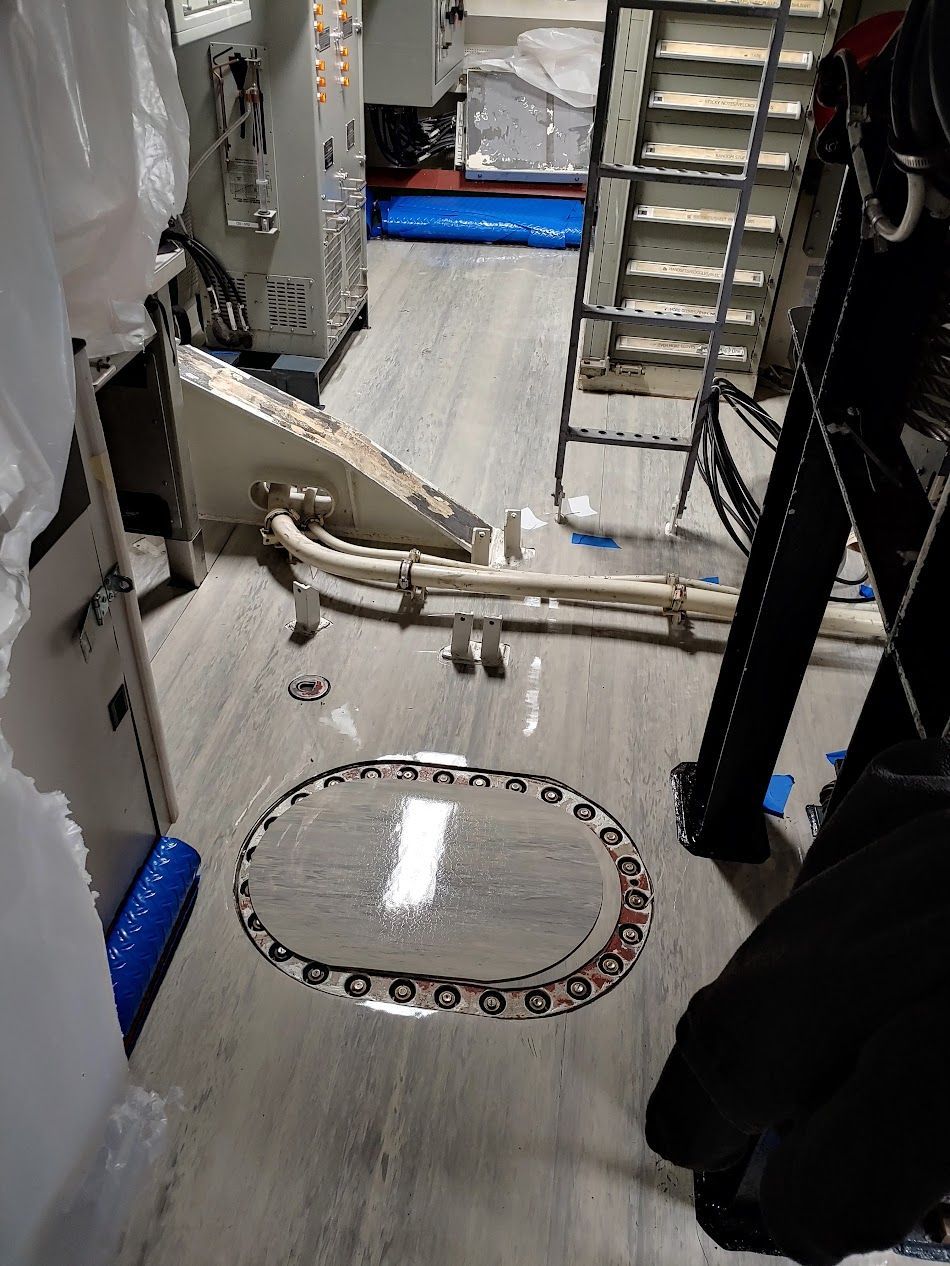 Interior view of a room with metal paneling, a ladder, and an oval hatch. Gray flooring.