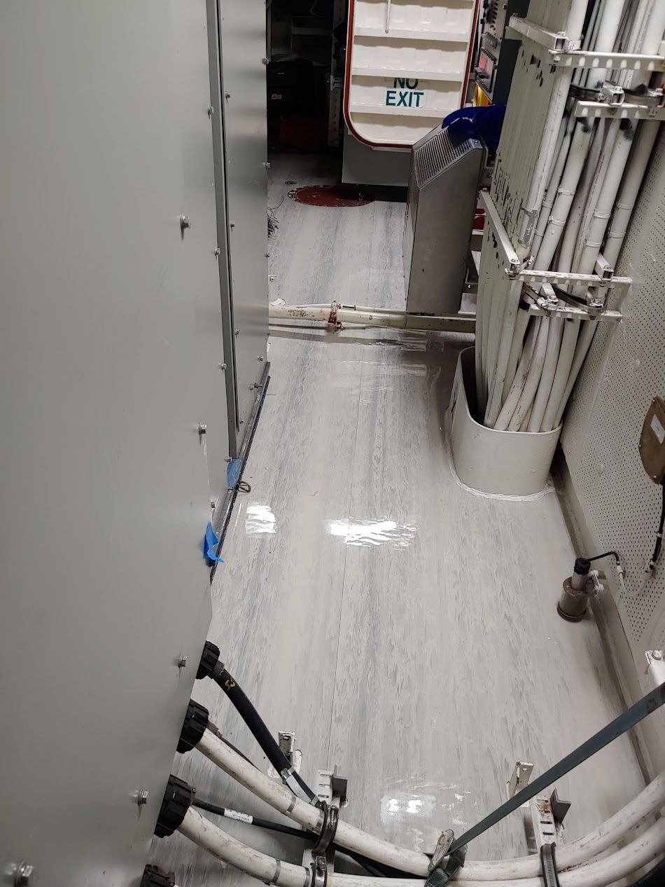 Narrow hallway with white floor, power cables, and electrical panels. Person visible in the background near an exit.