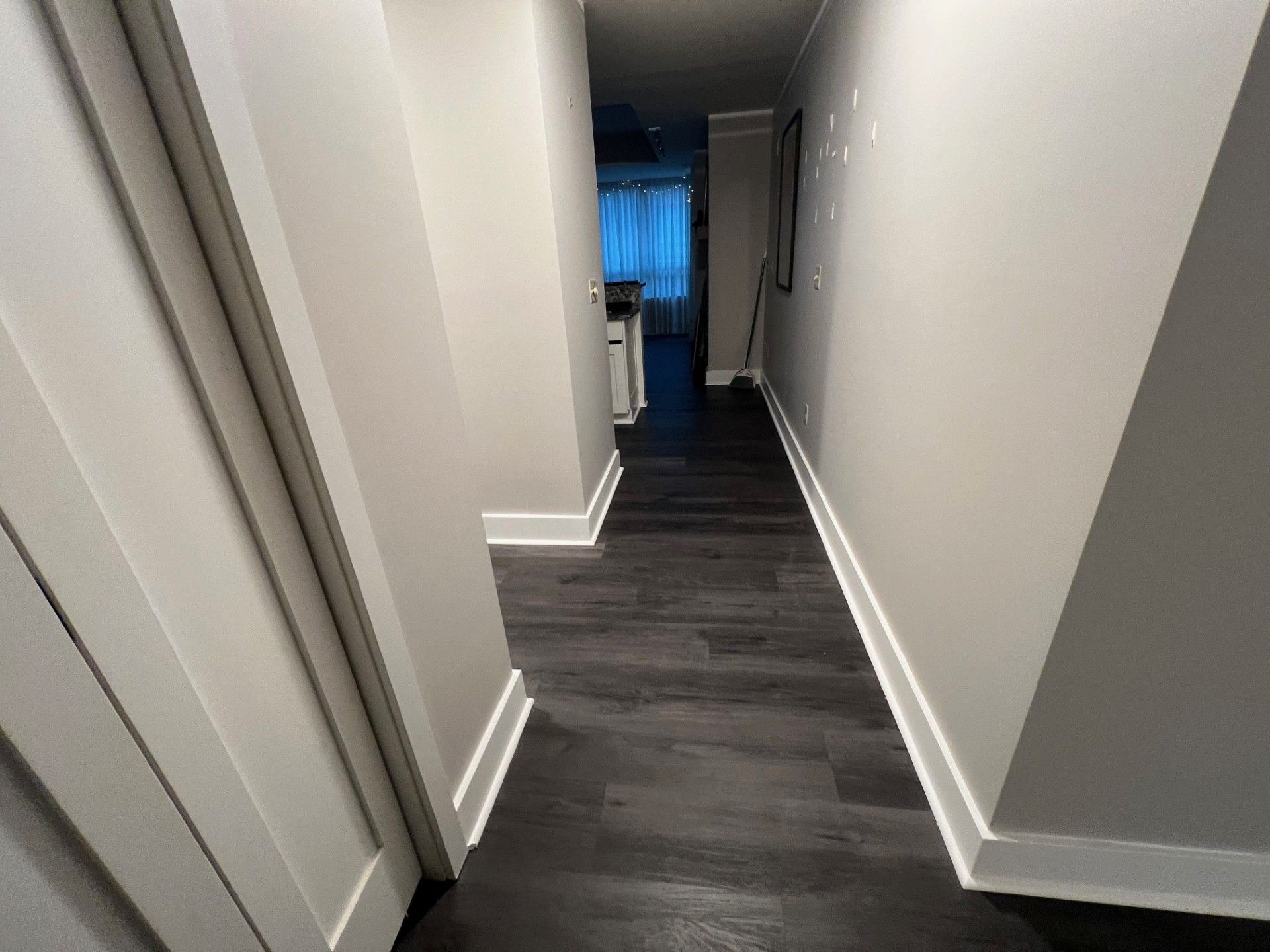 Narrow hallway with dark wood-look flooring and light gray walls, leading to a bright doorway.