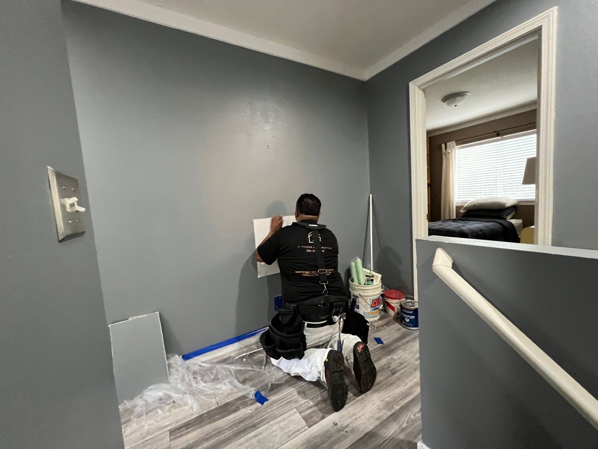 A person paints a wall gray in a hallway. They wear a black shirt, sitting on a drop cloth.