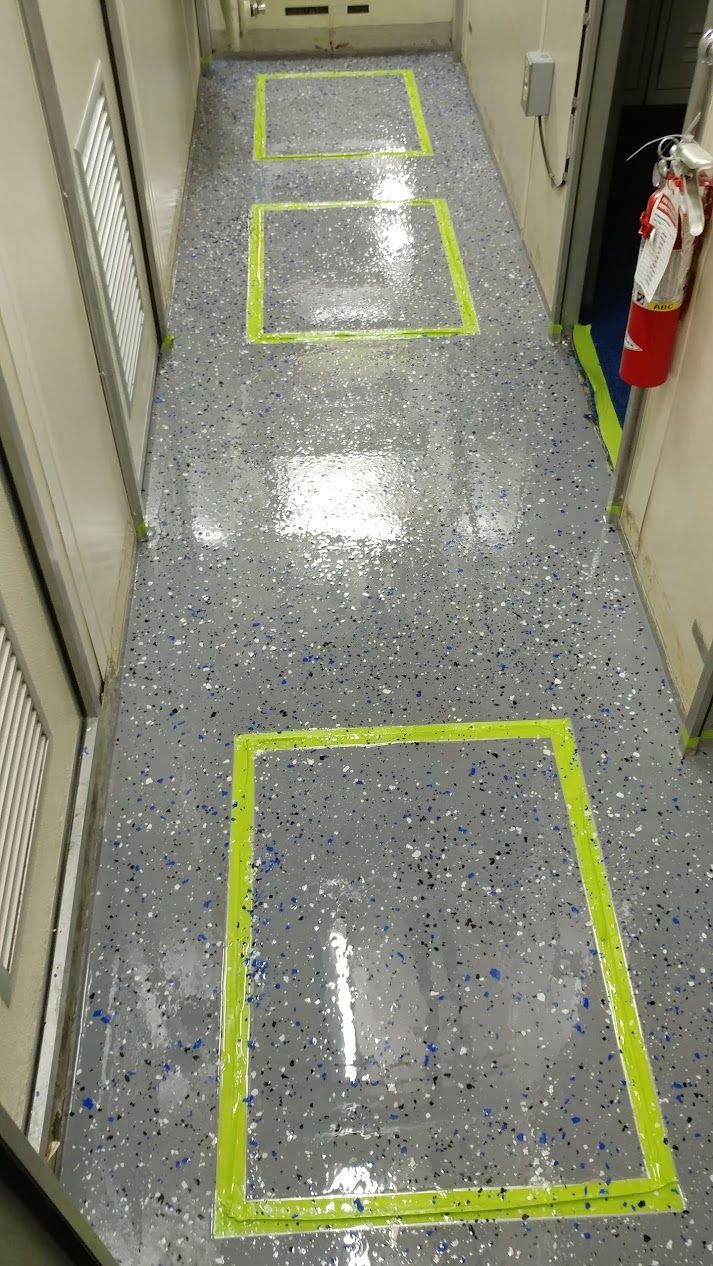 Narrow hallway with three yellow-taped squares on speckled gray floor. A fire extinguisher is visible on the right.